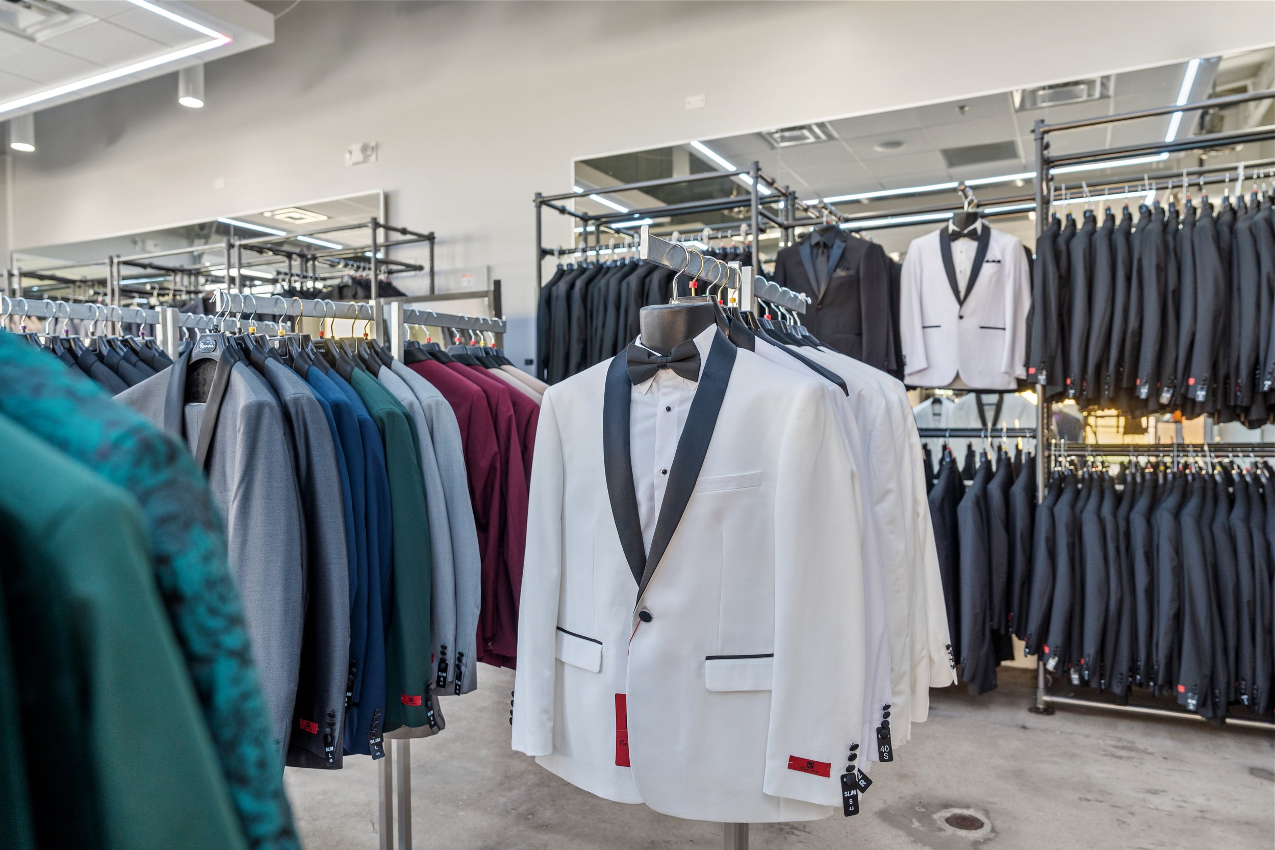 A display of men's suits and tuxedos on racks inside a clothing store, including a white tuxedo with a black lapel and bow tie.