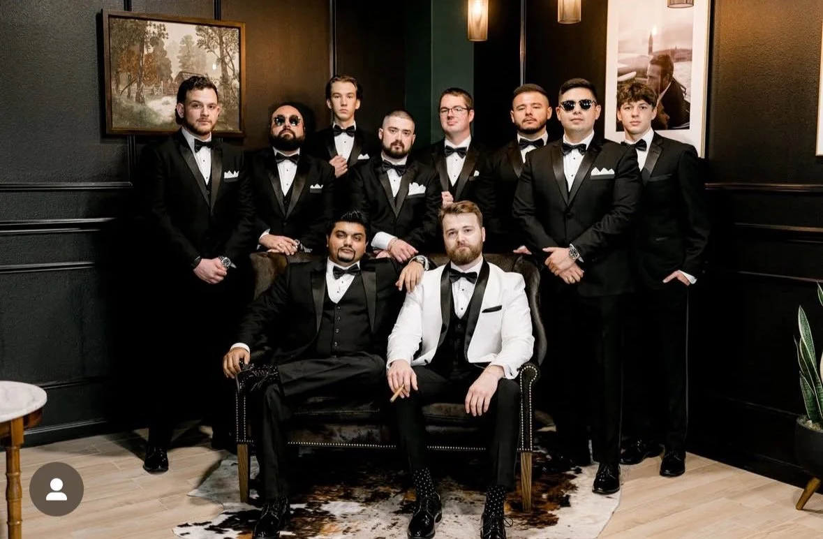 A group of eleven men in tuxedos, some with sunglasses, posing in a stylish, dimly lit room with dark walls, framed artwork, and a cowhide rug, some sitting and others standing behind them.
