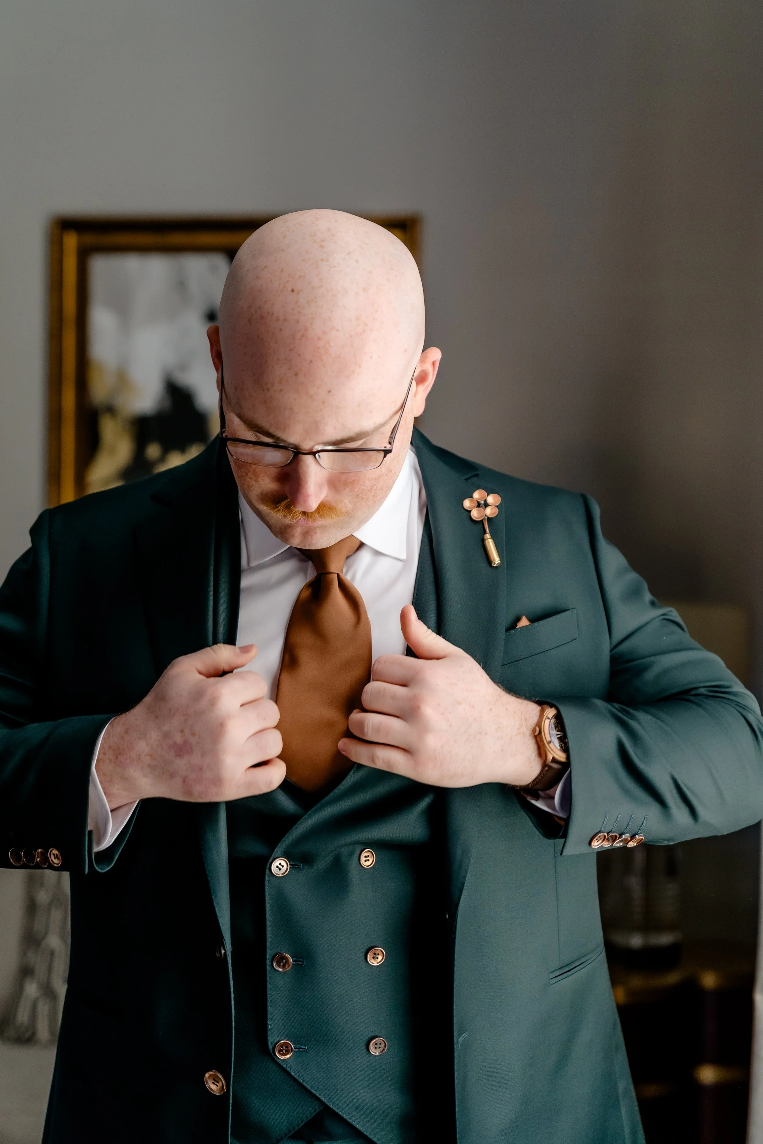 A man with glasses adjusts his brown tie while wearing a dark green suit and a brooch on his lapel.