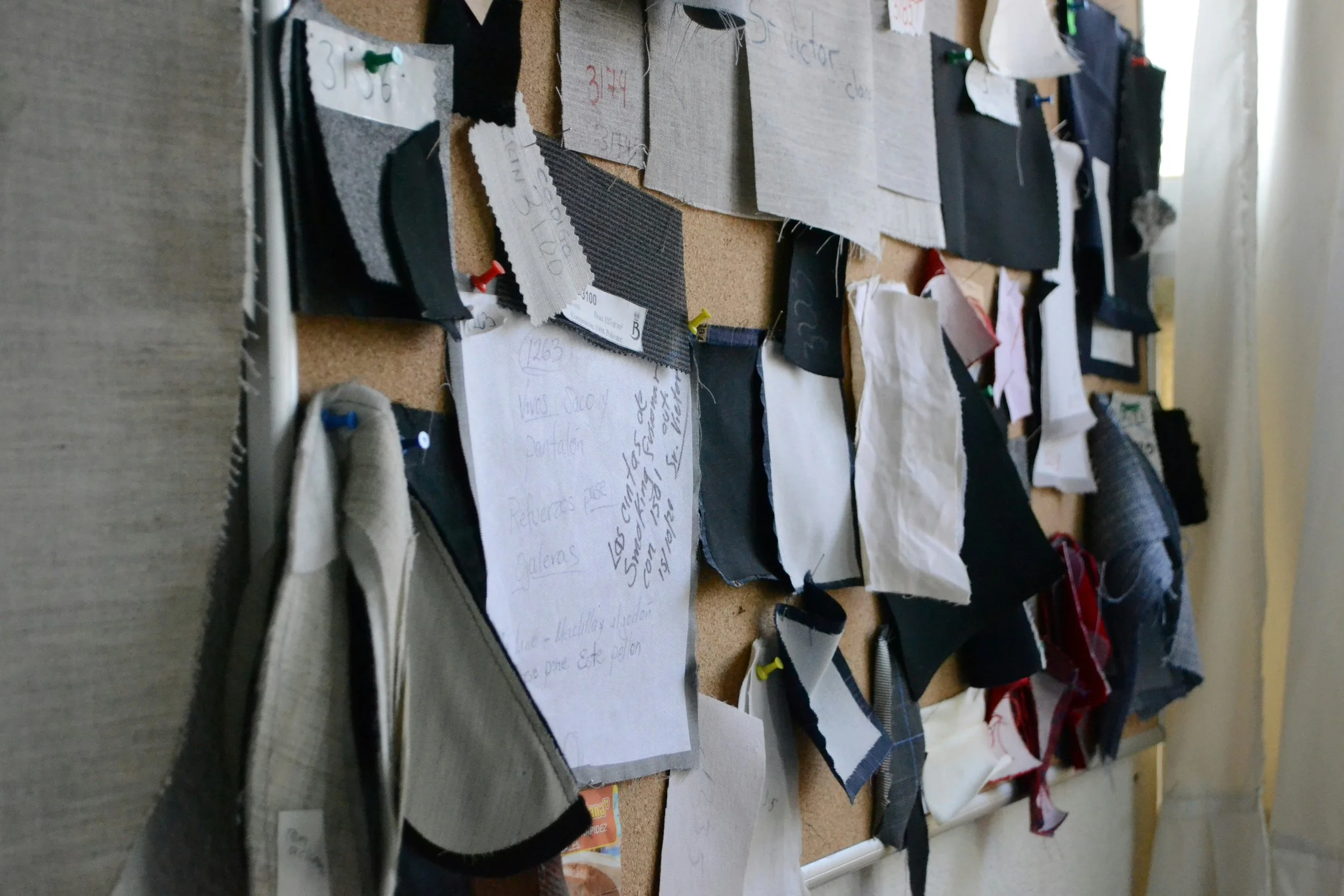 A corkboard filled with fabric swatches, notes, and sketches pinned with colored thumbtacks, used for sewing or fashion design. Some fabric pieces are taped or pinned and handwritten notes are visible.