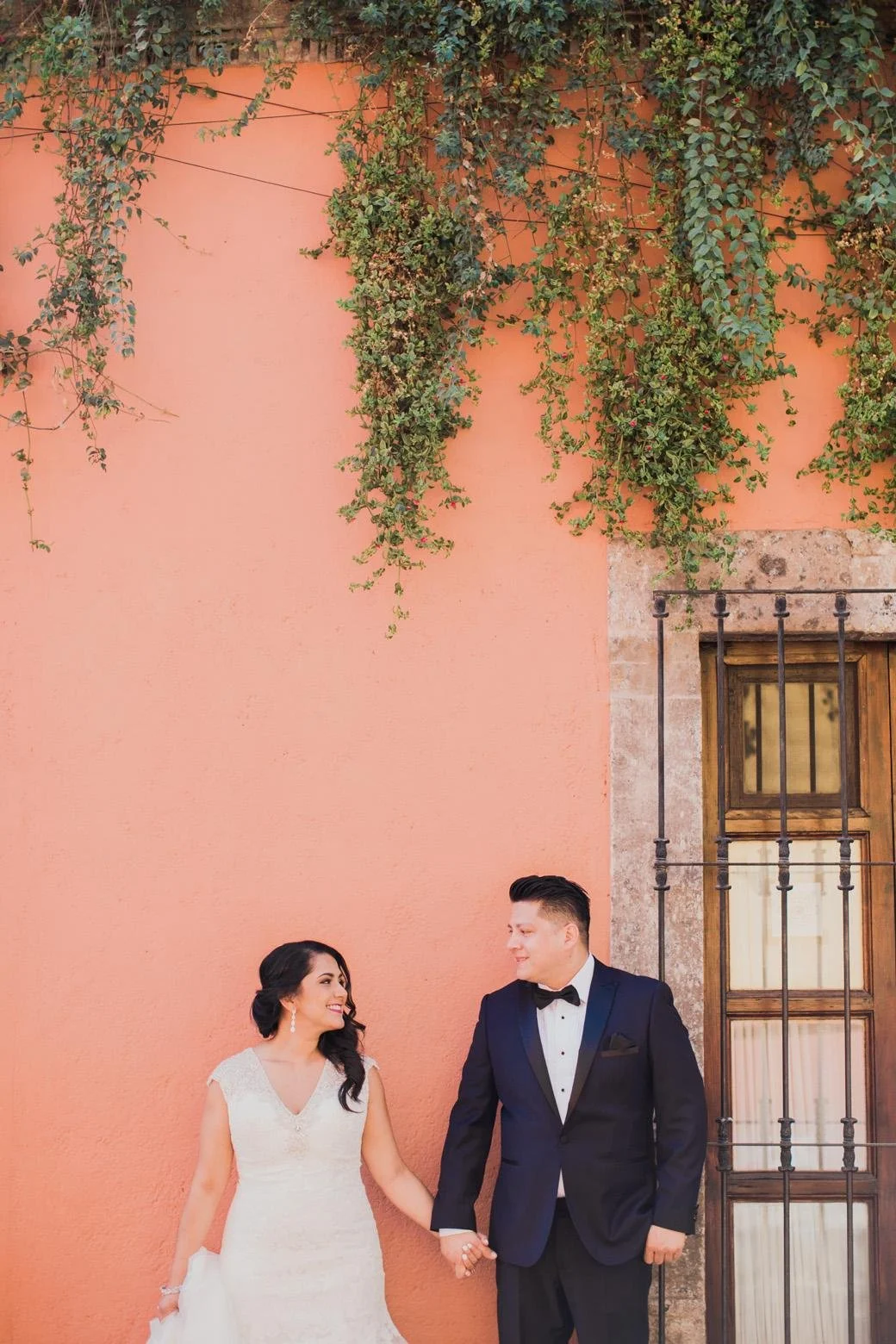 A bride and groom holding hands, smiling at each other, standing against a peach-colored wall with green vines and a wooden window.