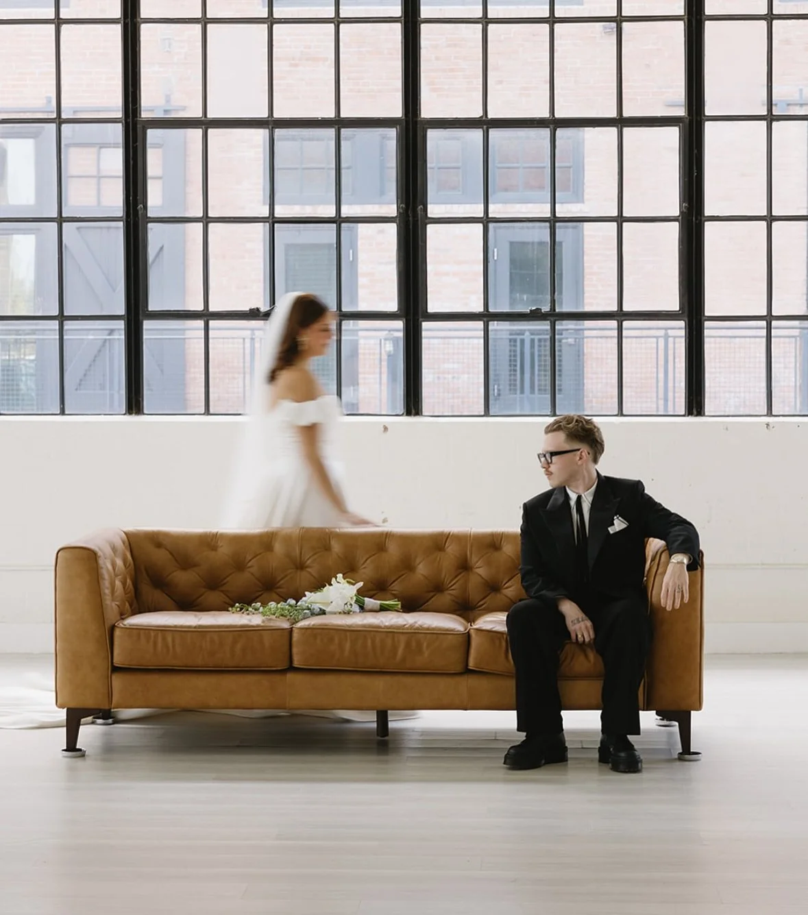 A woman in a white dress walking past a man in a black suit sitting on a brown leather sofa in front of large industrial-style windows, with a wedding bouquet on the sofa.