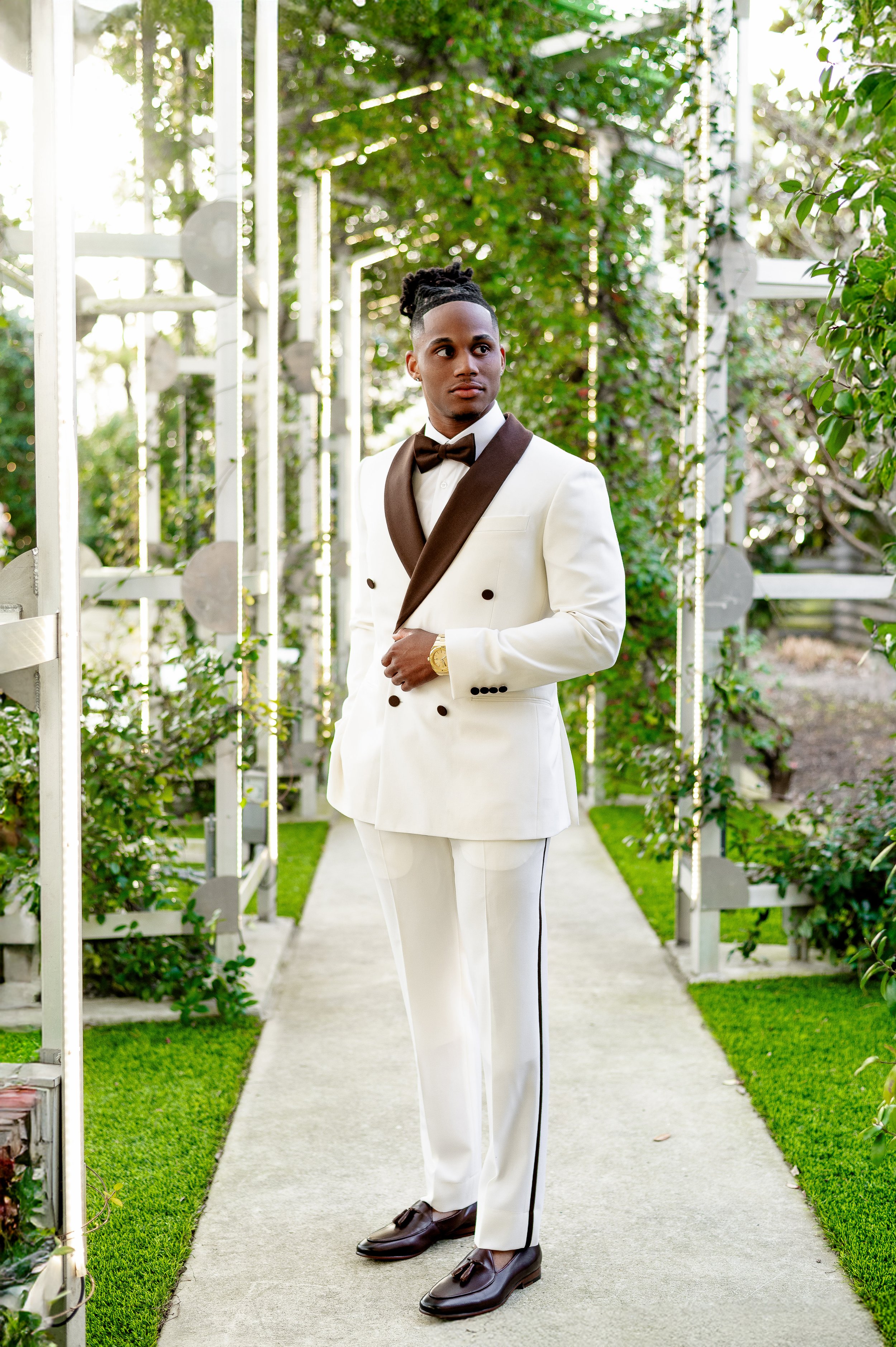 A man in a white tuxedo with a brown bow tie and brown lapel, standing on a garden pathway with greenery and structures in the background.