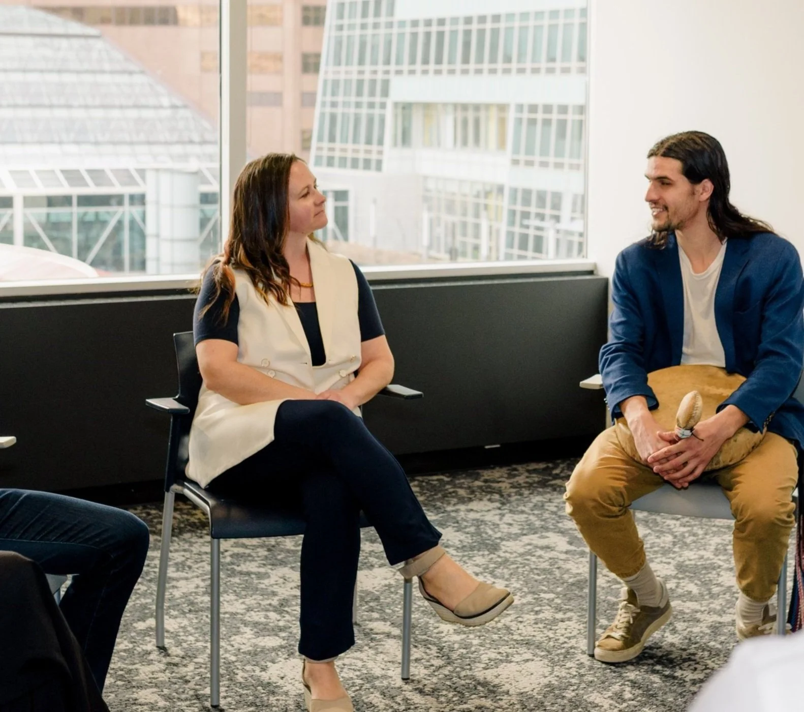 Two people sitting in a room having a conversation, with large city buildings visible through the window behind them.