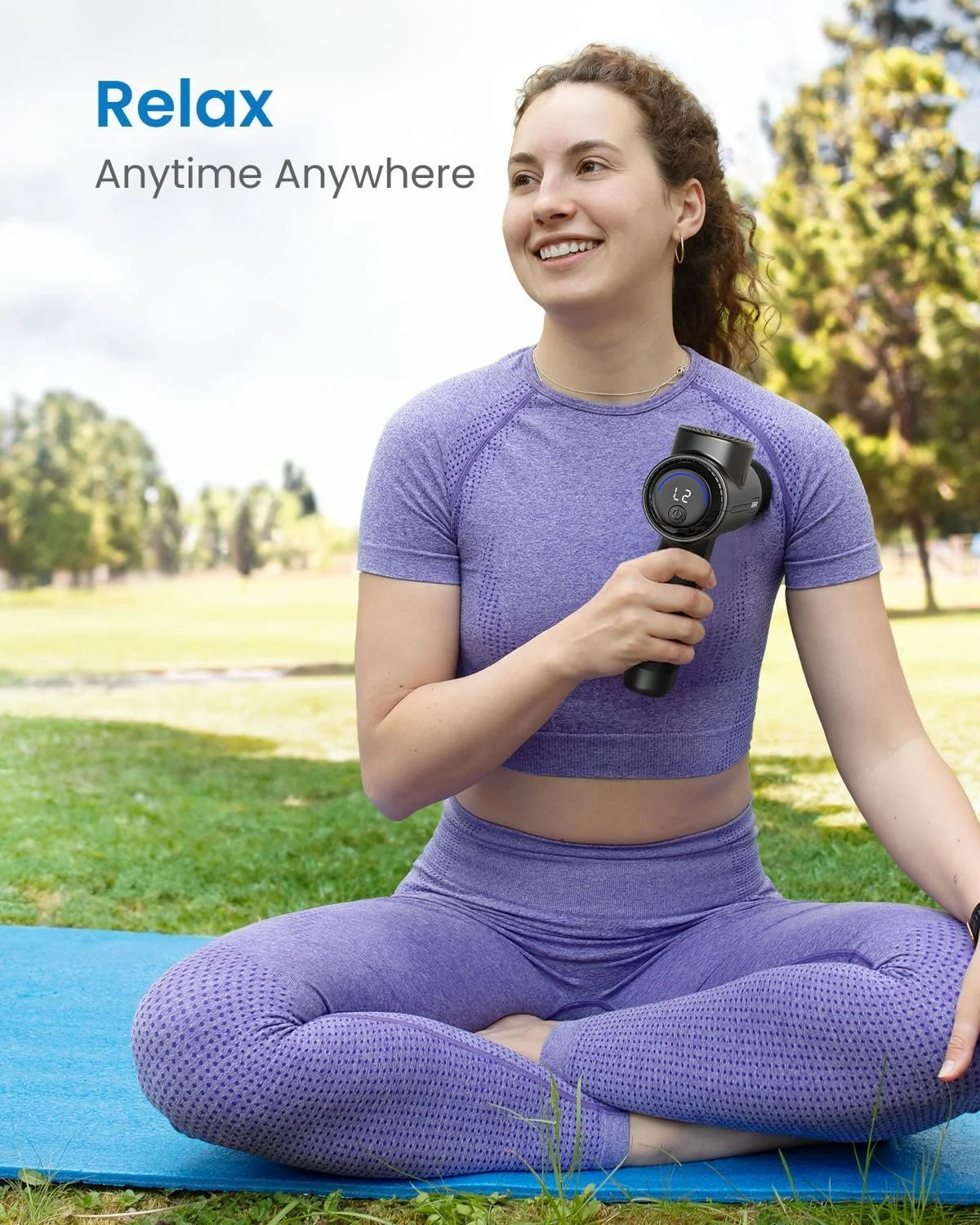 A woman in purple workout clothes sitting cross-legged on a blue yoga mat outdoors, holding a handheld massage gun, smiling, with trees and a grassy field in the background.