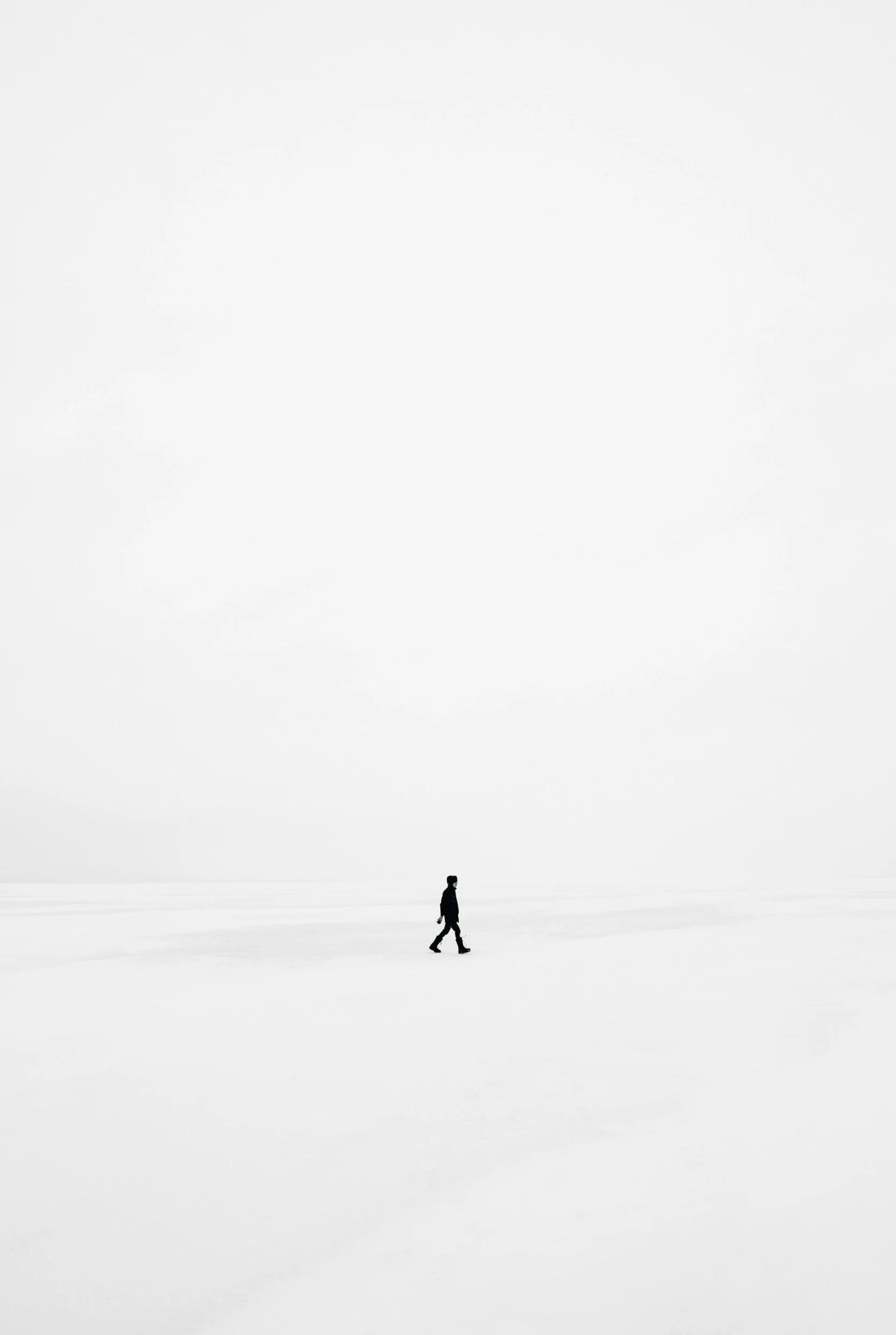 A person walking alone in a vast, snow-covered landscape with a mostly white and overcast sky.