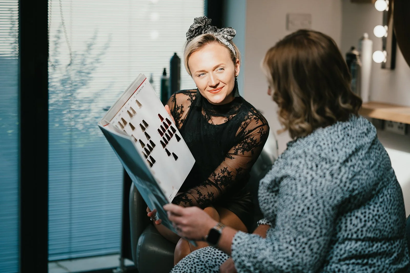 Two women sitting and talking in a salon, one woman holding a magazine or catalog showing hair color options.