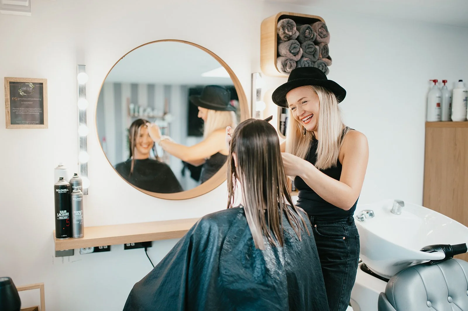 A hairstylist with a black hat smiling as she styles a woman's wet hair in a salon. The salon has a round mirror, hair products, towels, and a sink.