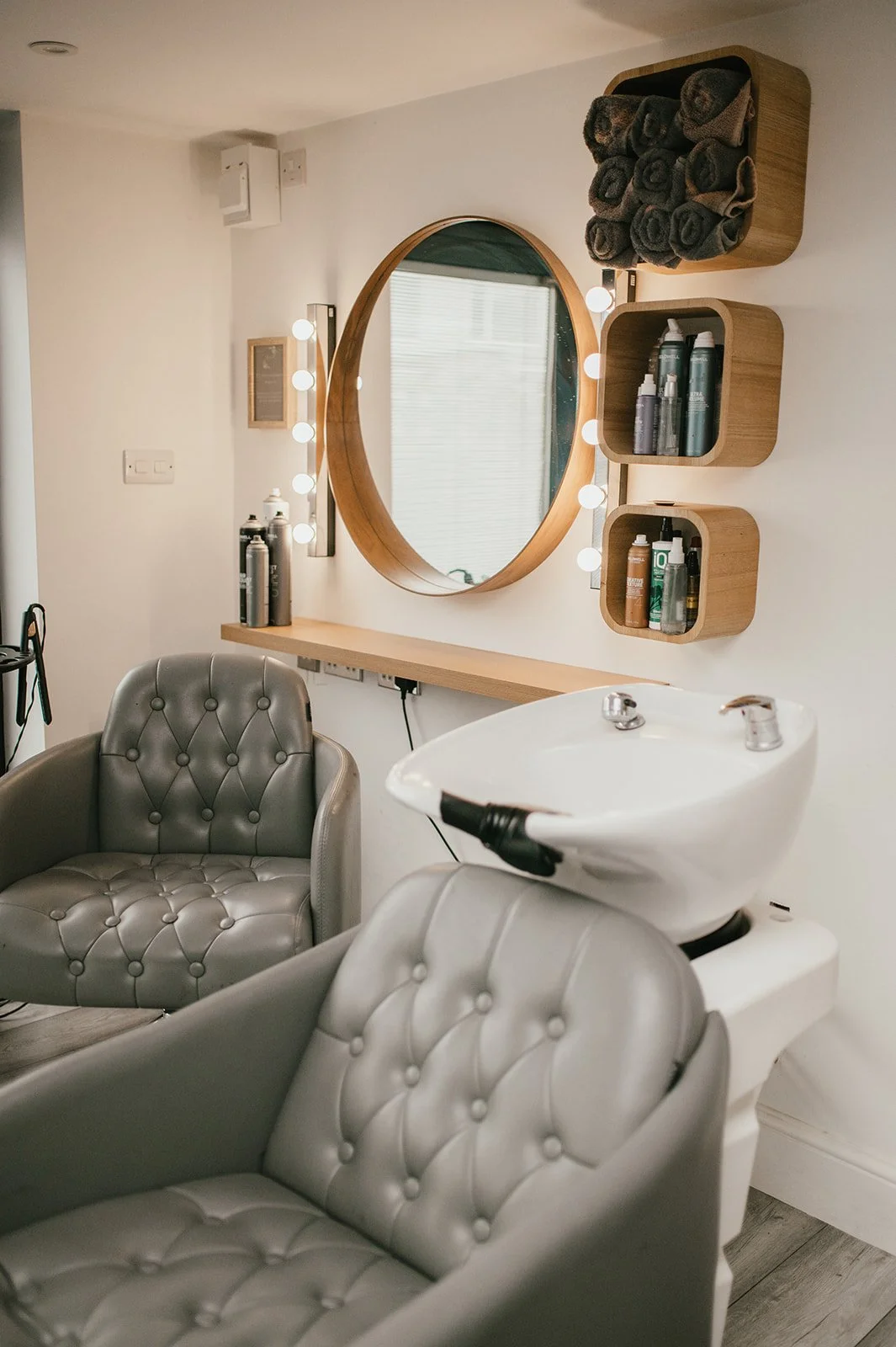 A hair salon with two light grey tufted chairs, a hair washing sink, a large wooden-framed round mirror, wooden shelves with hair products, and a wooden shelf with rolled towels.