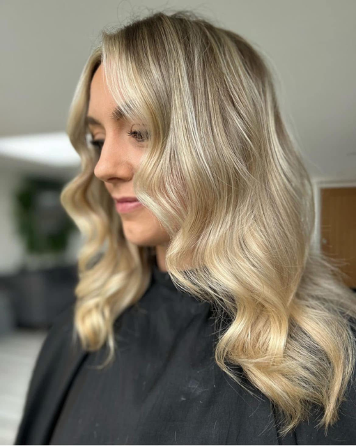A woman with wavy blonde hair styled in loose waves, wearing a black cape, in a salon setting.