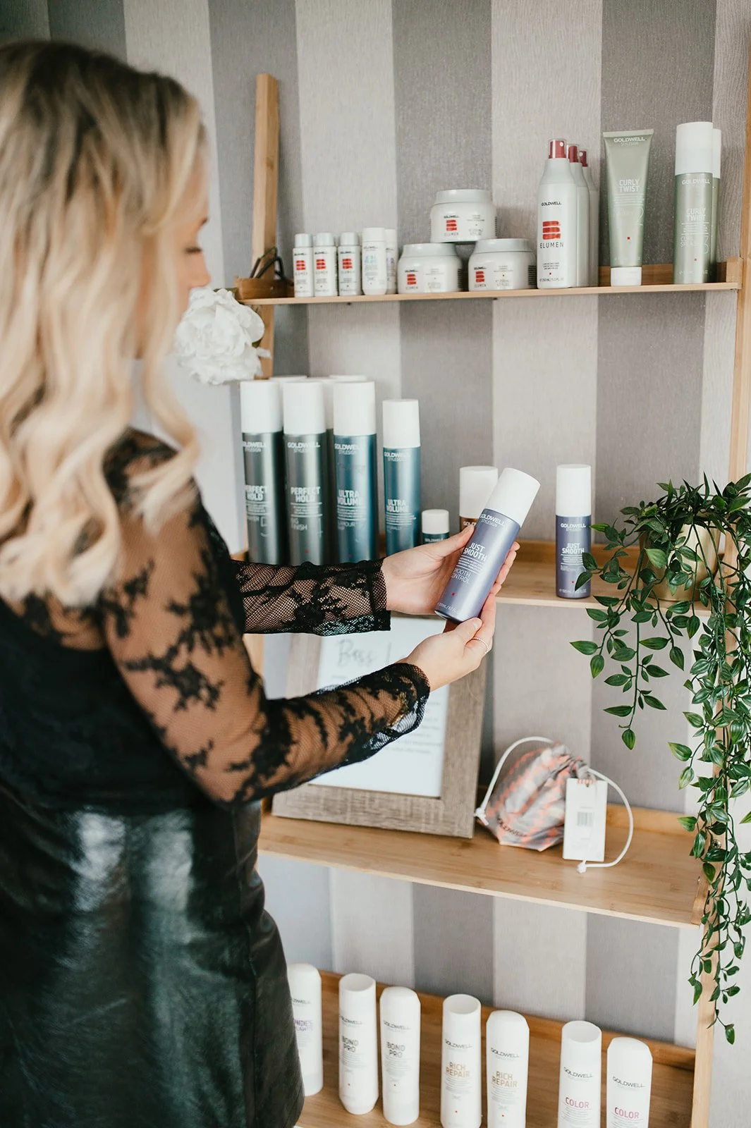 A woman with blonde hair in a black lace top is holding a can of Goldwell hair spray, with shelves of hair care products in the background.