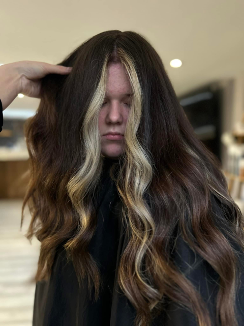 Young woman with long, wavy brown hair with blonde highlights, sitting in a hair salon with her eyes closed.