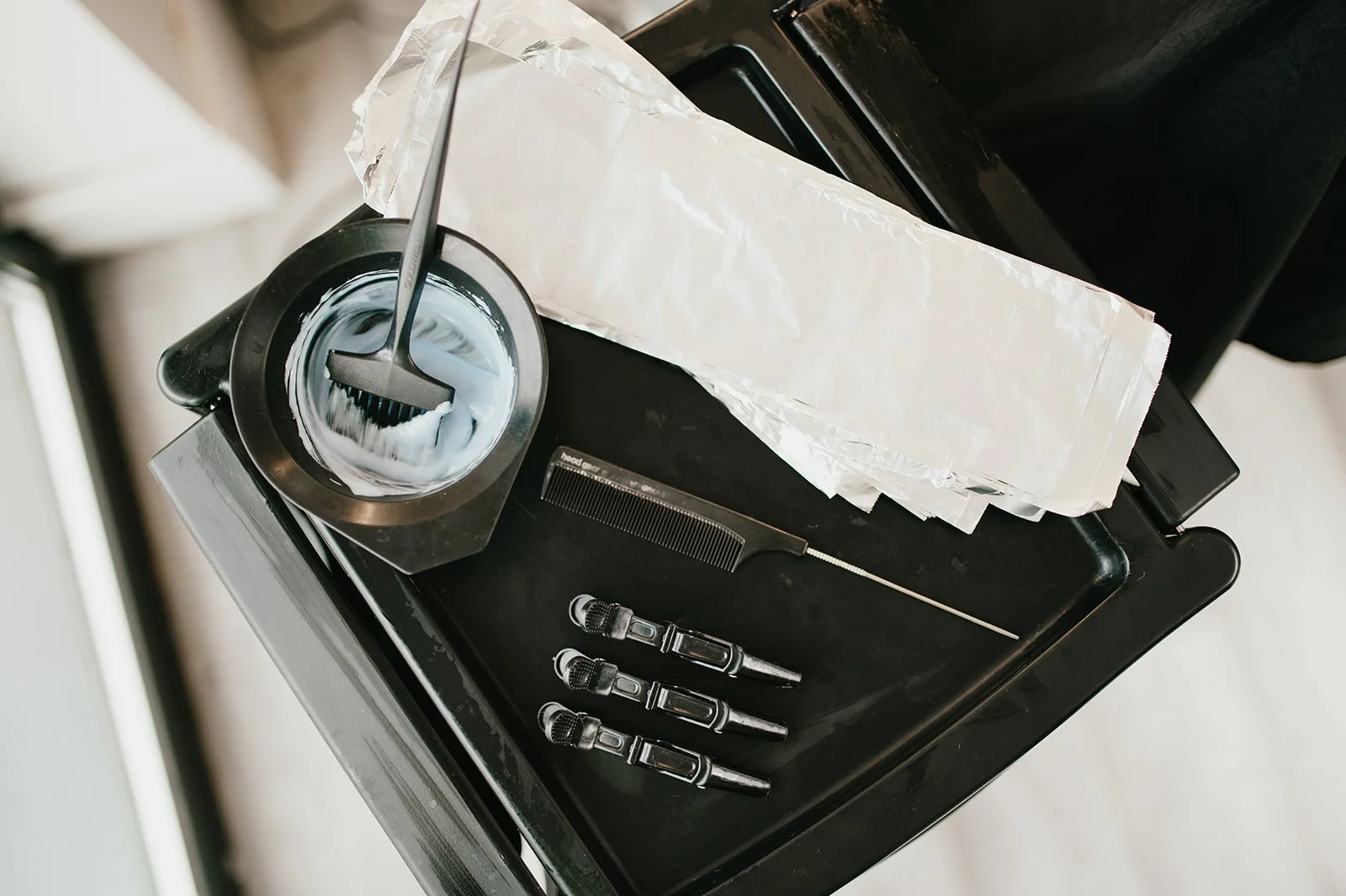 Hairdressing tools on a black cart, including foil, a brush in a bowl, black clips, and a long thin metal tool.