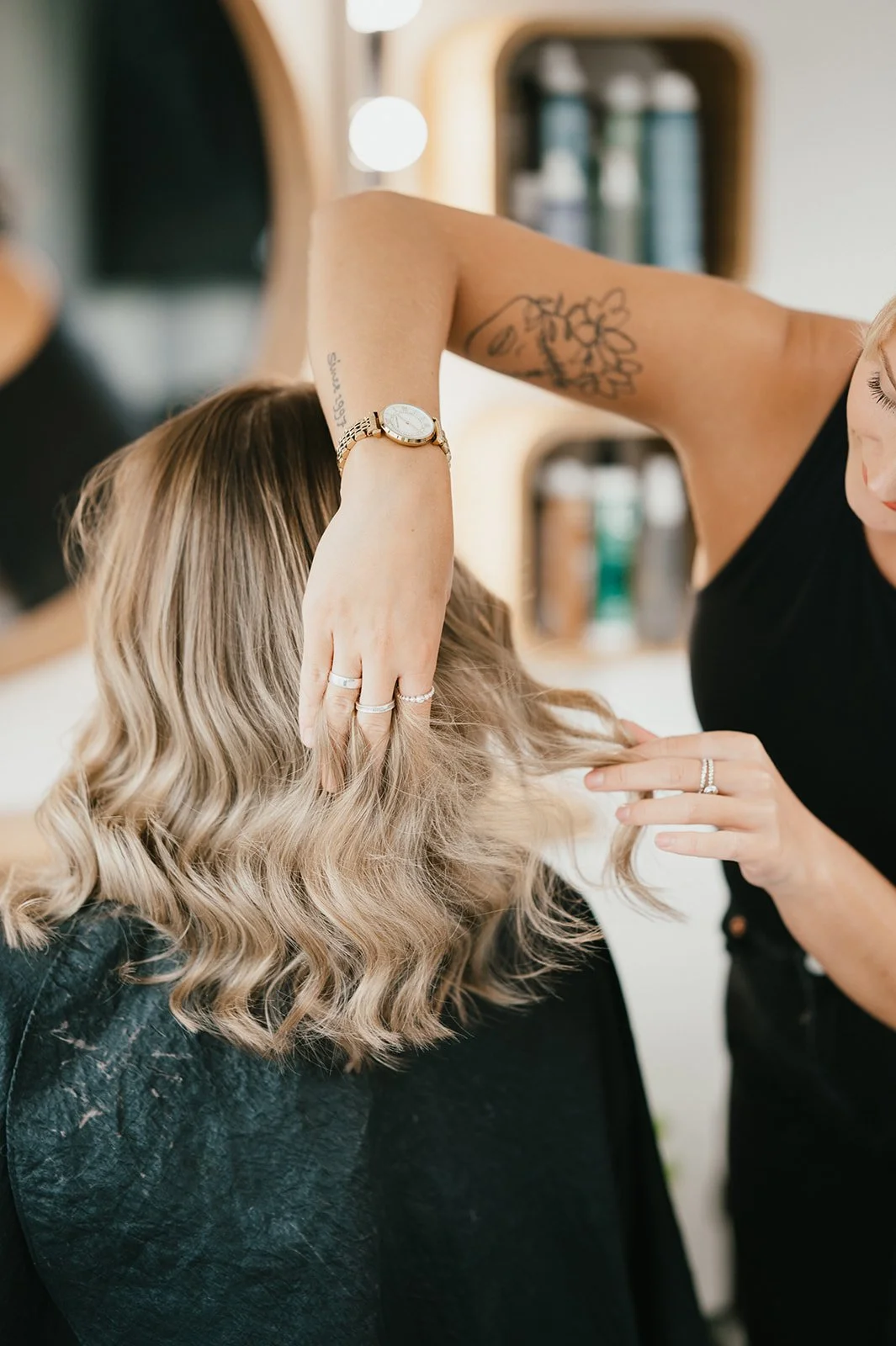 A hairstylist working on a woman's blonde, wavy hair in a salon, wearing rings and a watch.