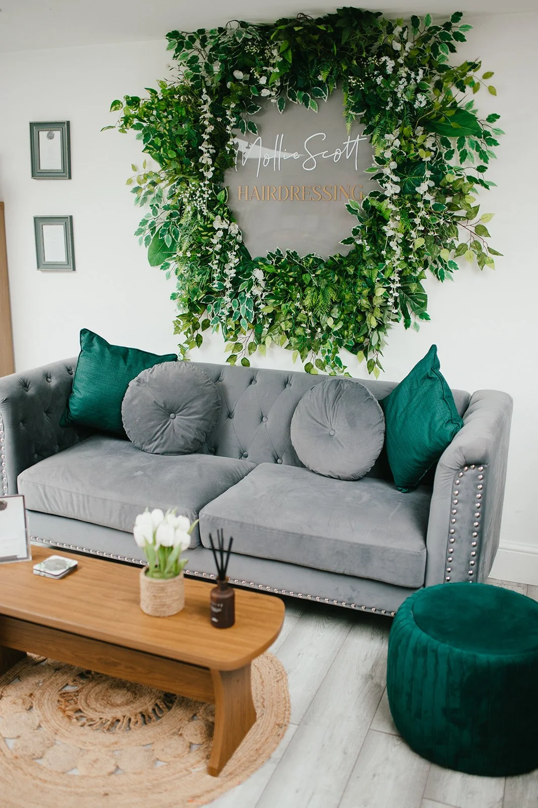 Waiting area with gray tufted sofa, green and gray pillows, wooden coffee table with flowers and reed diffuser, circular carpet, green ottoman, large leafy green wreath with signage reading 'Hollie Scott Hairdressing' on the wall.