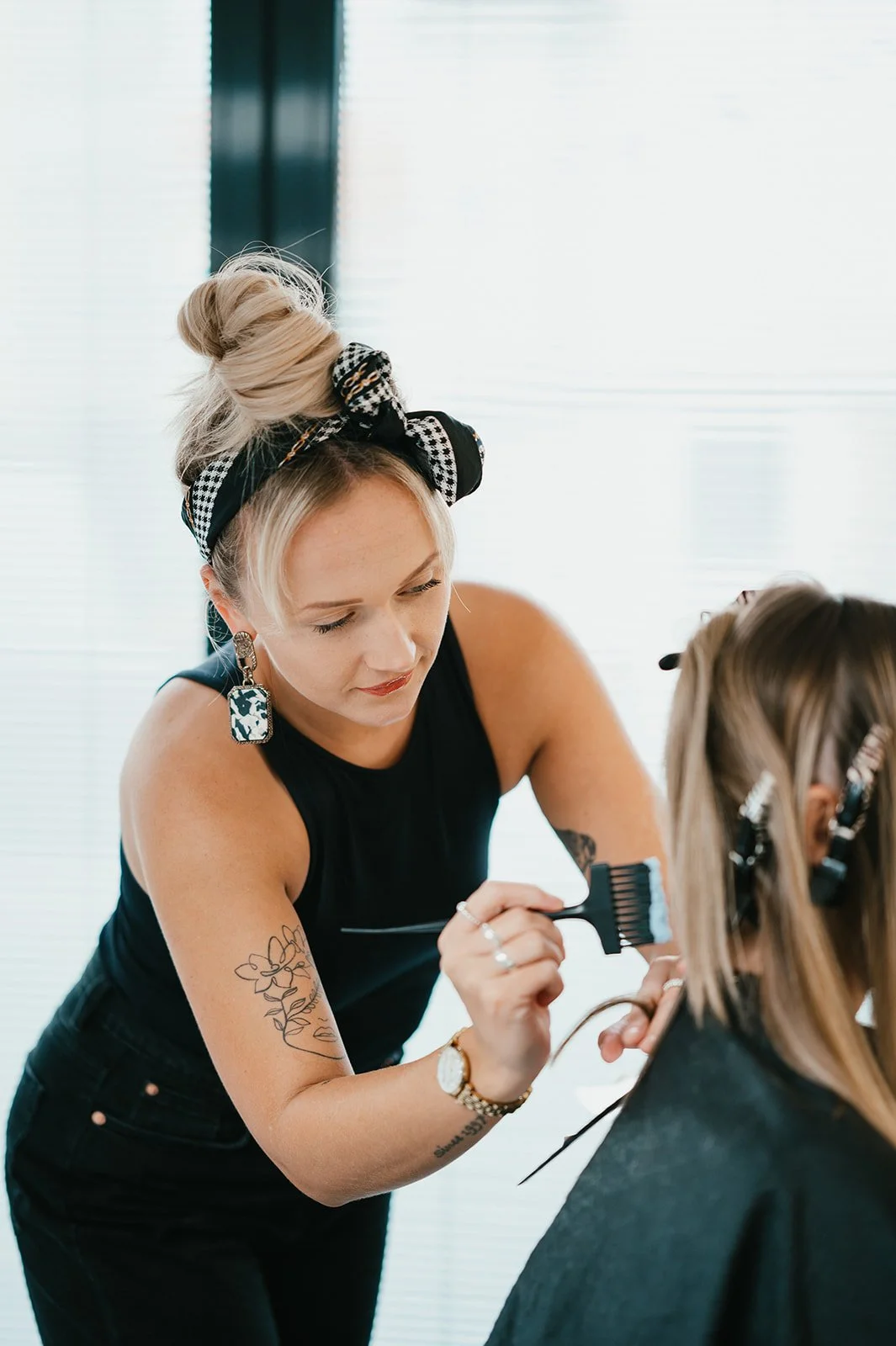 A female hairstylist with blonde hair in a bun, wearing a black sleeveless top and checkered headband, is dyeing a client's hair.