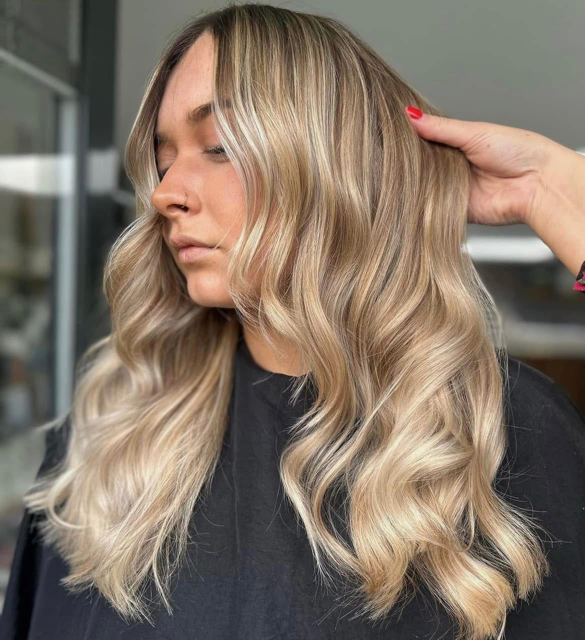 Woman with long, wavy blonde hair and light highlights, seated in a hair salon.