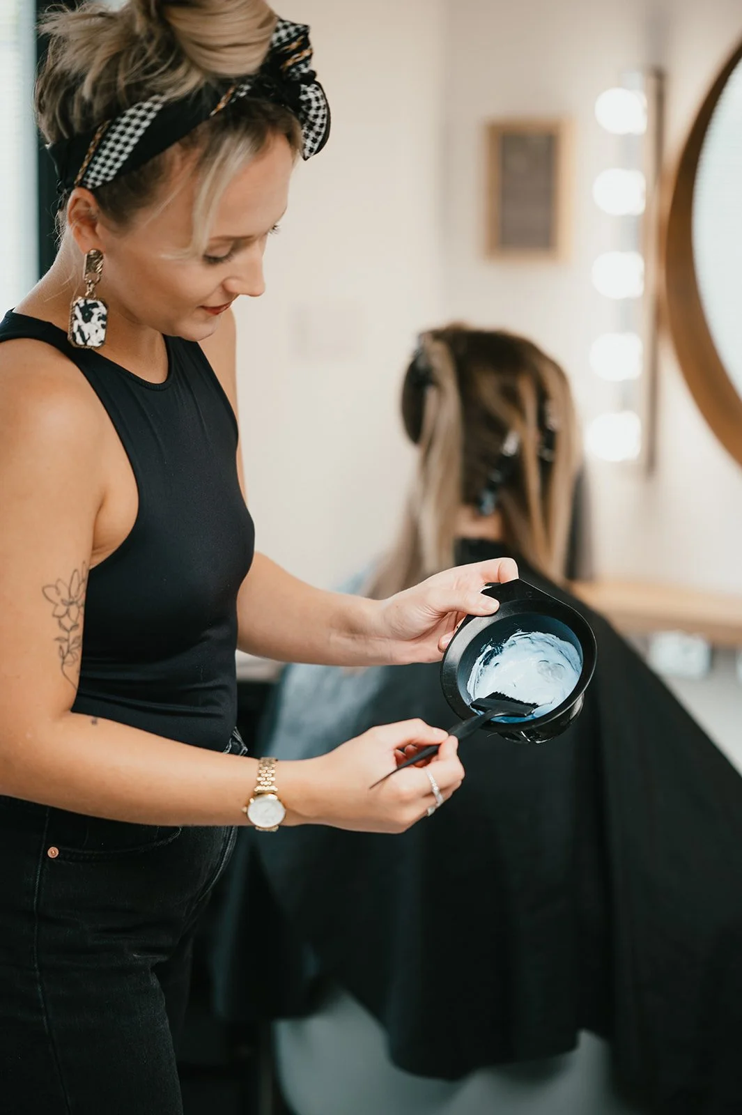 A woman with wavy blonde hair, wearing a black tank top, a black and white checkered headband, large earrings, and a gold watch, is standing at a mirror in a hair salon. She is holding a black plastic bowl with a white cream or hair product and a sma