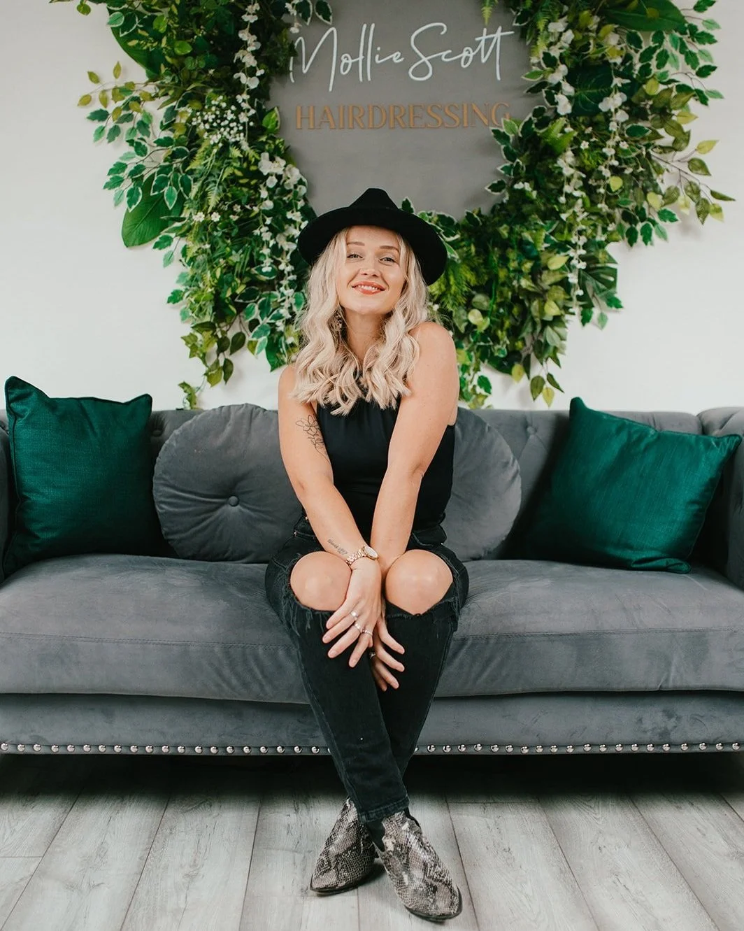 A woman with blonde wavy hair wearing a black dress, black ripped jeans, and snakeskin ankle boots, sitting on a gray couch with green pillows, in front of a leafy green wall with a sign reading 'Mollie Scott Hairdressing,' smiling at the camera.