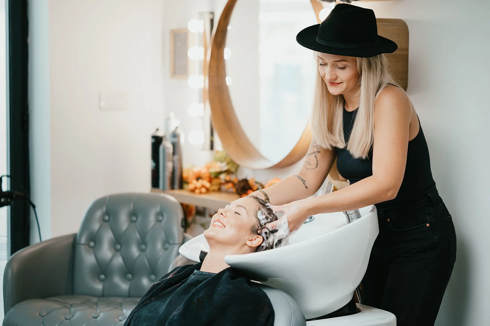 A hairdresser washing a woman's hair at a salon sink, with a mirror, salon supplies, and decorative items in the background.
