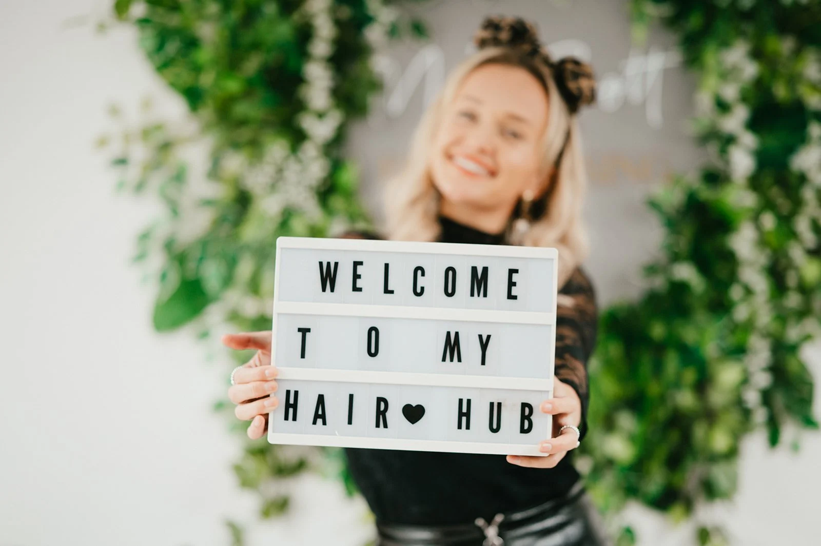 A woman with blonde hair and black hair accessories holding a sign that says 'WELCOME TO MY HAIR HUB', blurred background of green plants.