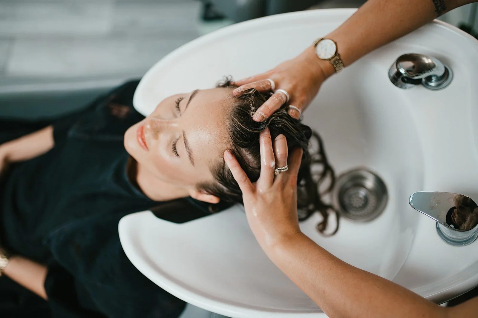 Woman receiving a hair wash at a salon sink with eyes closed, while stylist's hands gently wash her hair.