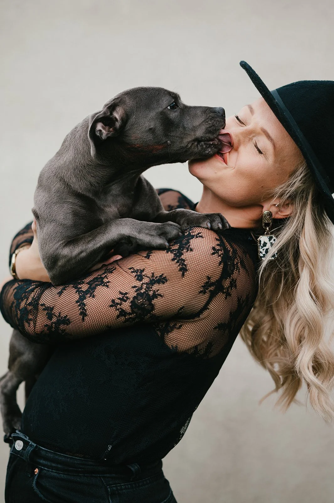 A woman with long, curly blonde hair wearing a black lace top, earrings, and a black wide-brimmed hat, holding a small gray puppy and kissing it on the face.