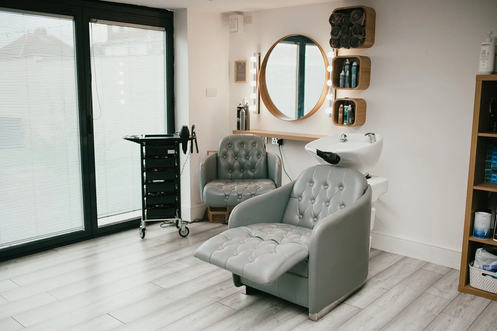 Empty hair salon with salon chairs, mirror, sink, and storage shelves.