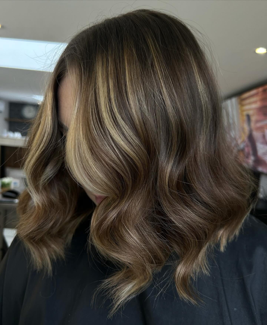 Woman with shoulder-length wavy hair with blonde highlights and darker roots, indoors in a salon or similar setting.