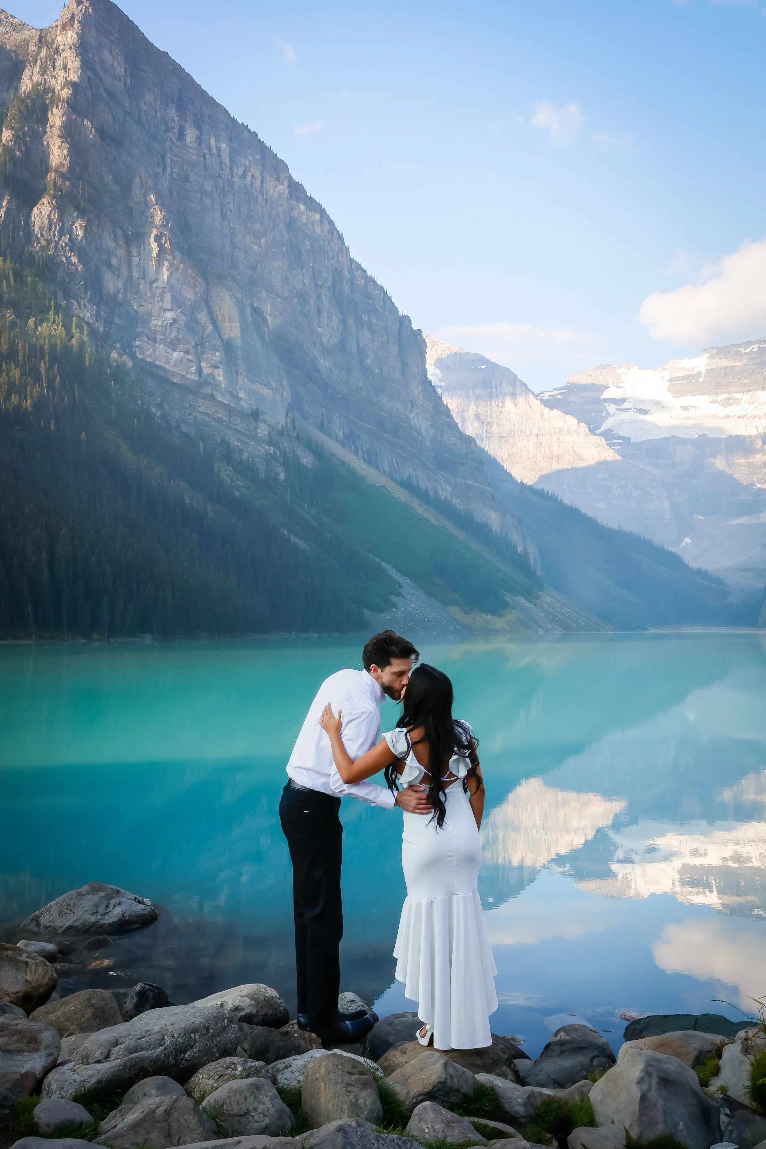 A couple dressed in white standing on rocks by a mountain lake, with mountains and a partly cloudy sky in the background.