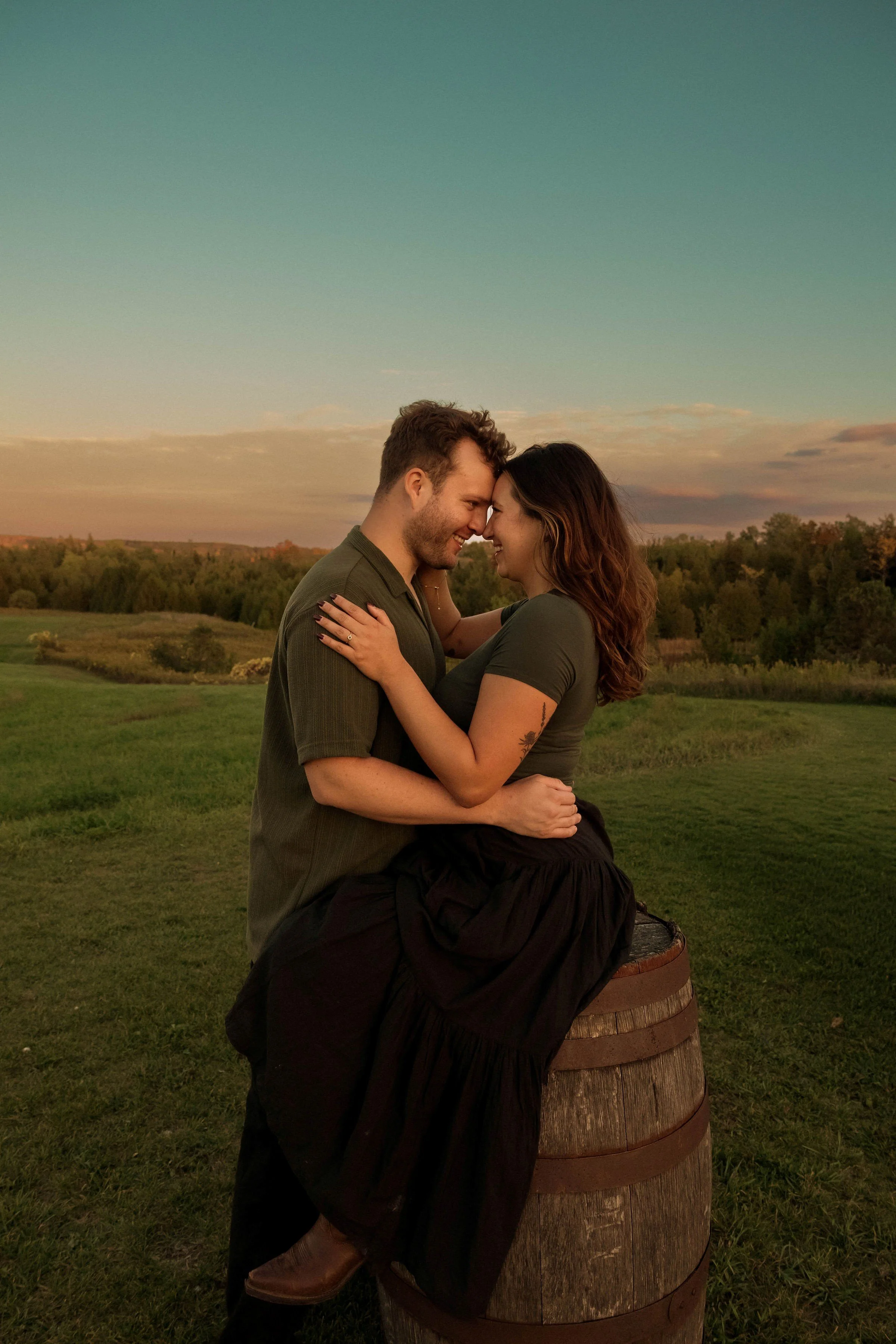 A couple standing outdoors at sunset, sharing an intimate moment with their foreheads touching, with a scenic landscape in the background.