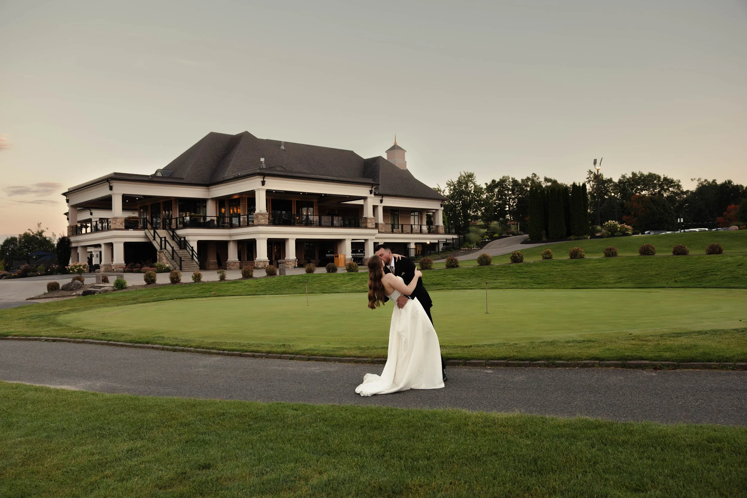 A bride and groom share a kiss on a golf course with a large two-story house in the background during sunset.