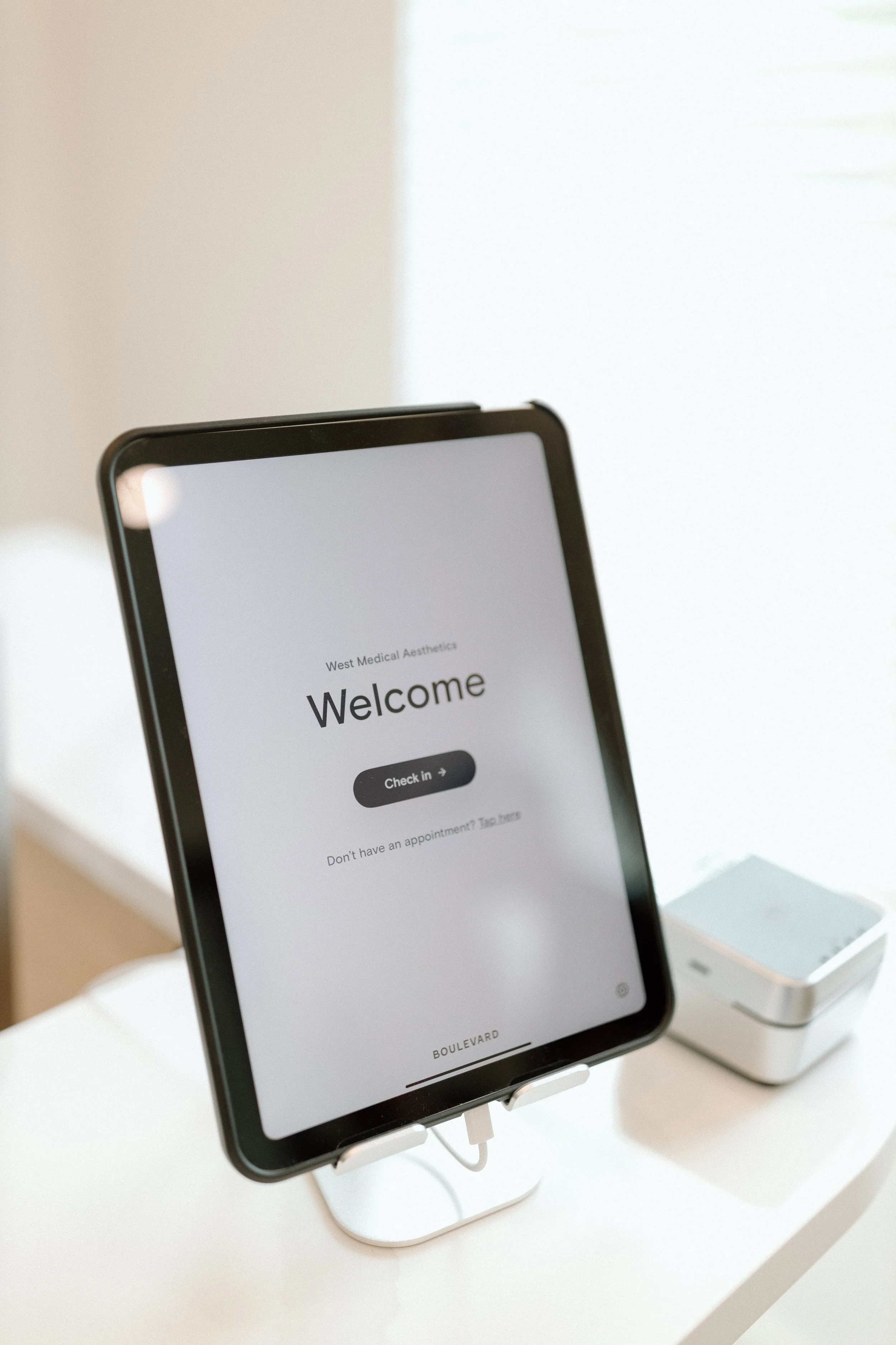 A tablet on a stand displaying a welcome message for West Medical Aesthetics with a check-in button, placed on a white surface with a small device next to it.