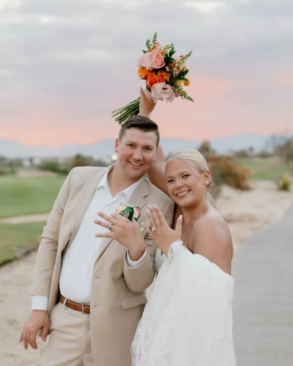 A happy bride and groom celebrate their wedding outdoors at sunset, showing off their wedding rings and bouquet, with the bride holding the groom’s shoulder and the groom holding a can of soda.