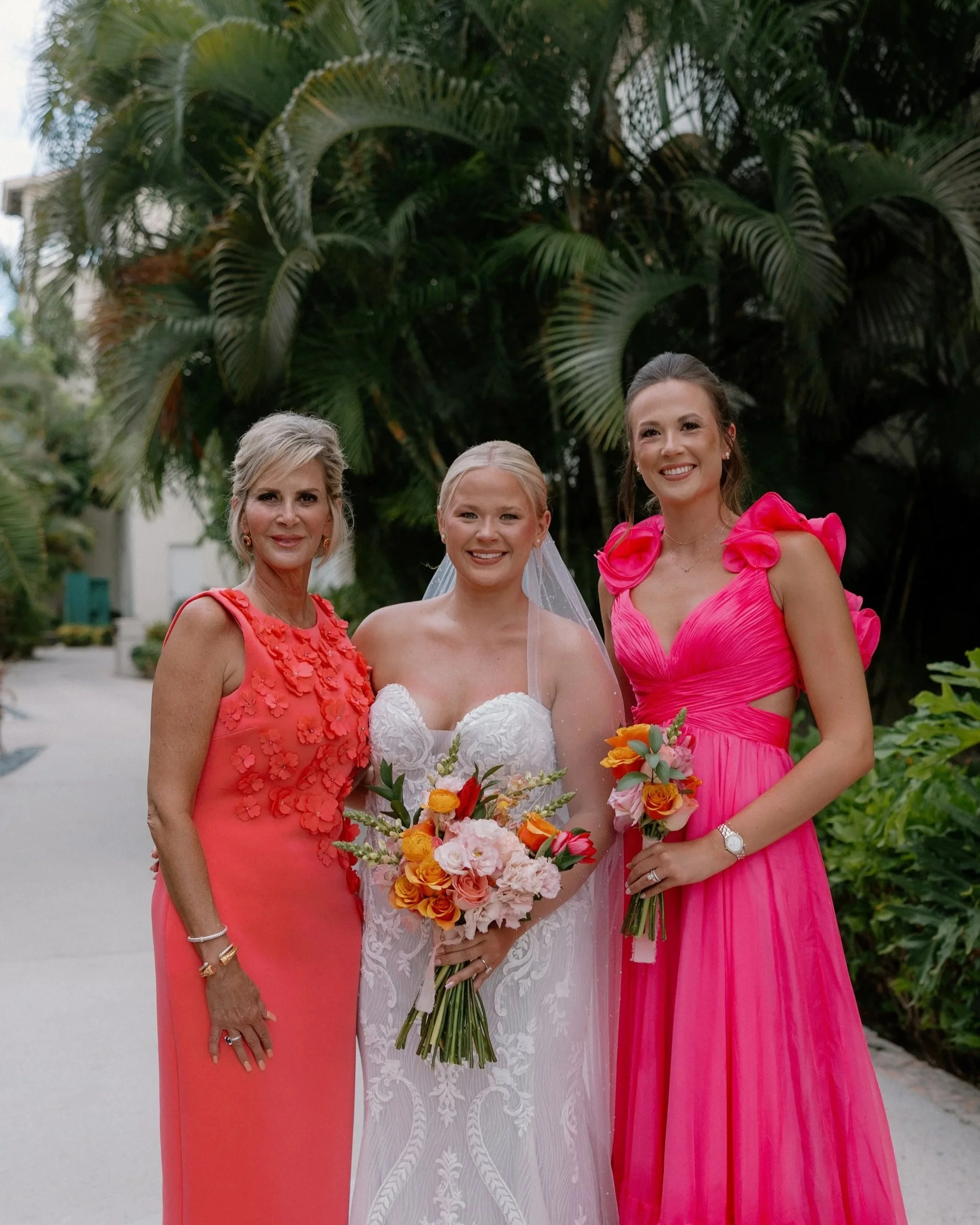Three women in colorful dresses smiling outdoors, with one woman in a white wedding gown holding a bouquet of flowers, and the others in pink dresses, standing in front of lush green tropical plants.