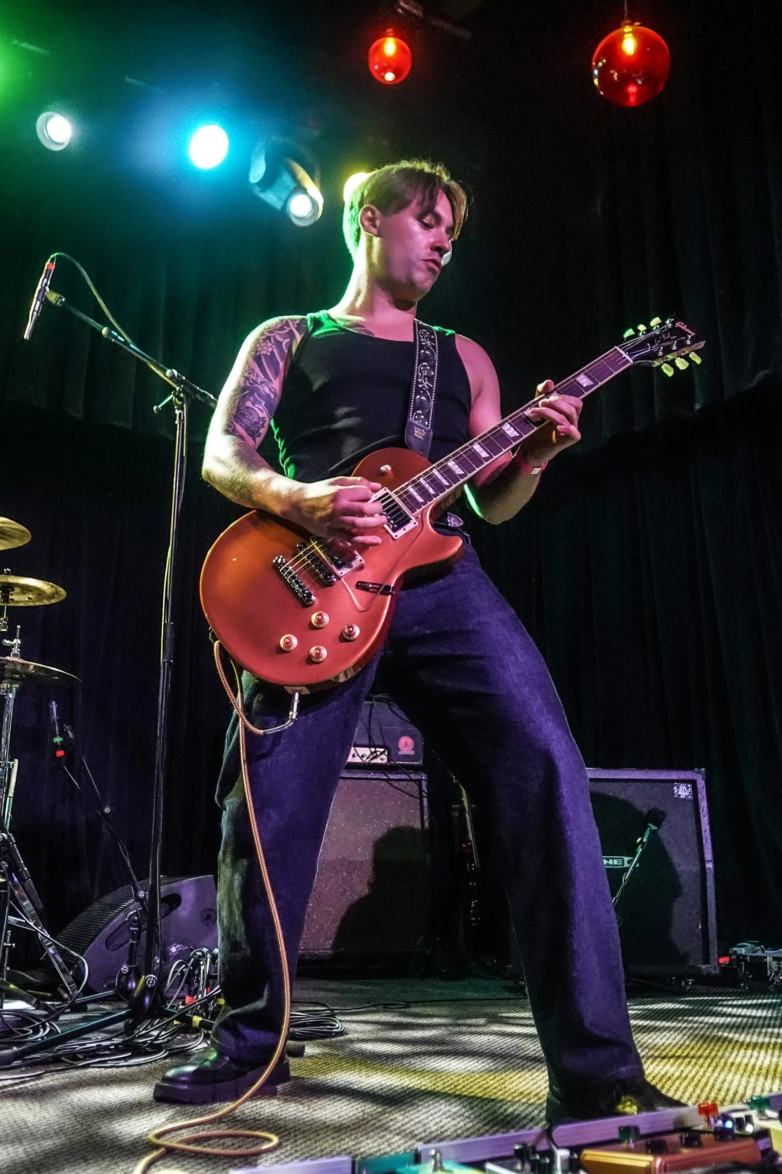 A musician in a black sleeveless shirt and jeans playing an electric guitar on stage with colored lighting and equipment around.