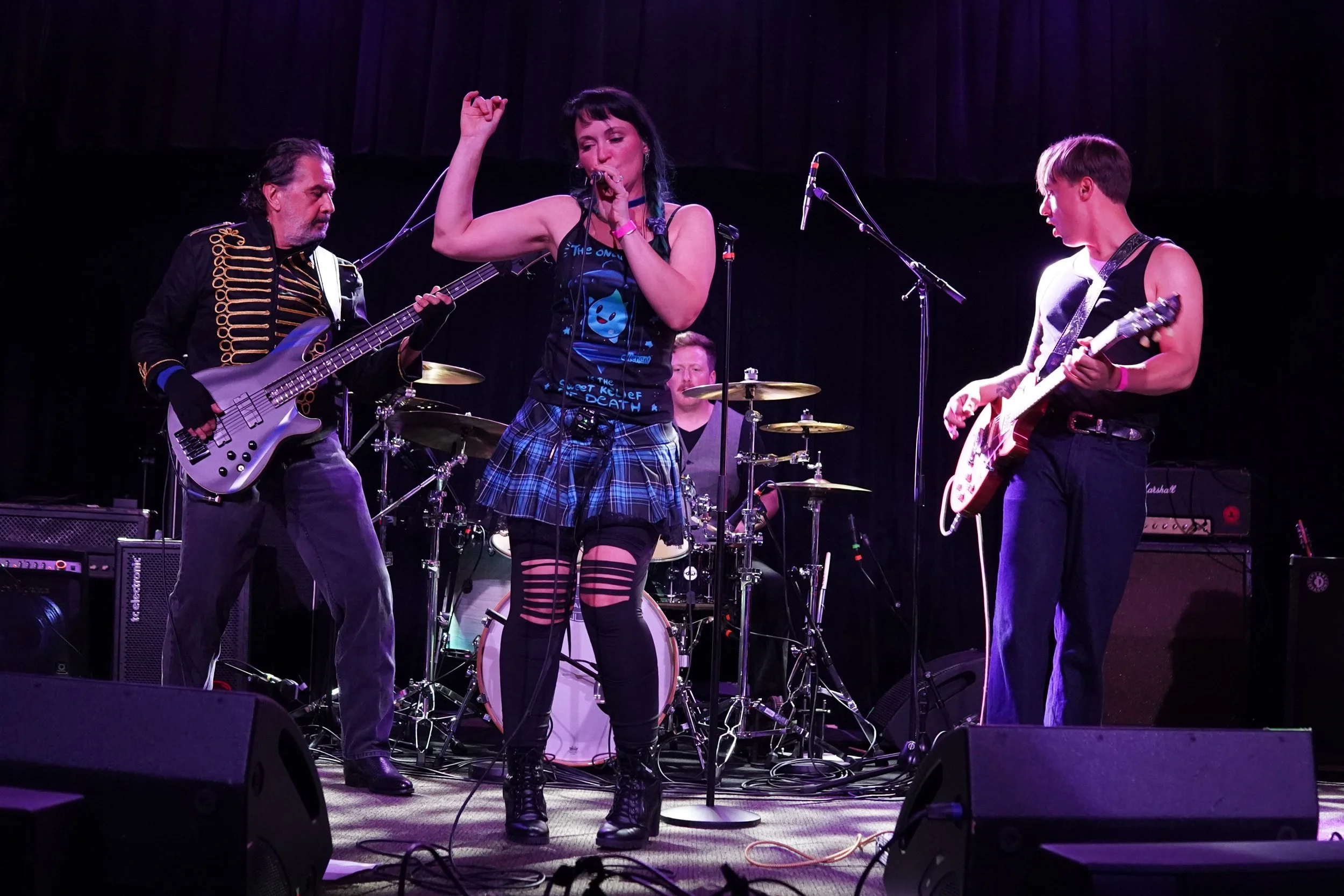A band performing on stage with a female singer in the center, flanked by a guitarist and a bassist, with a drum set in the background.
