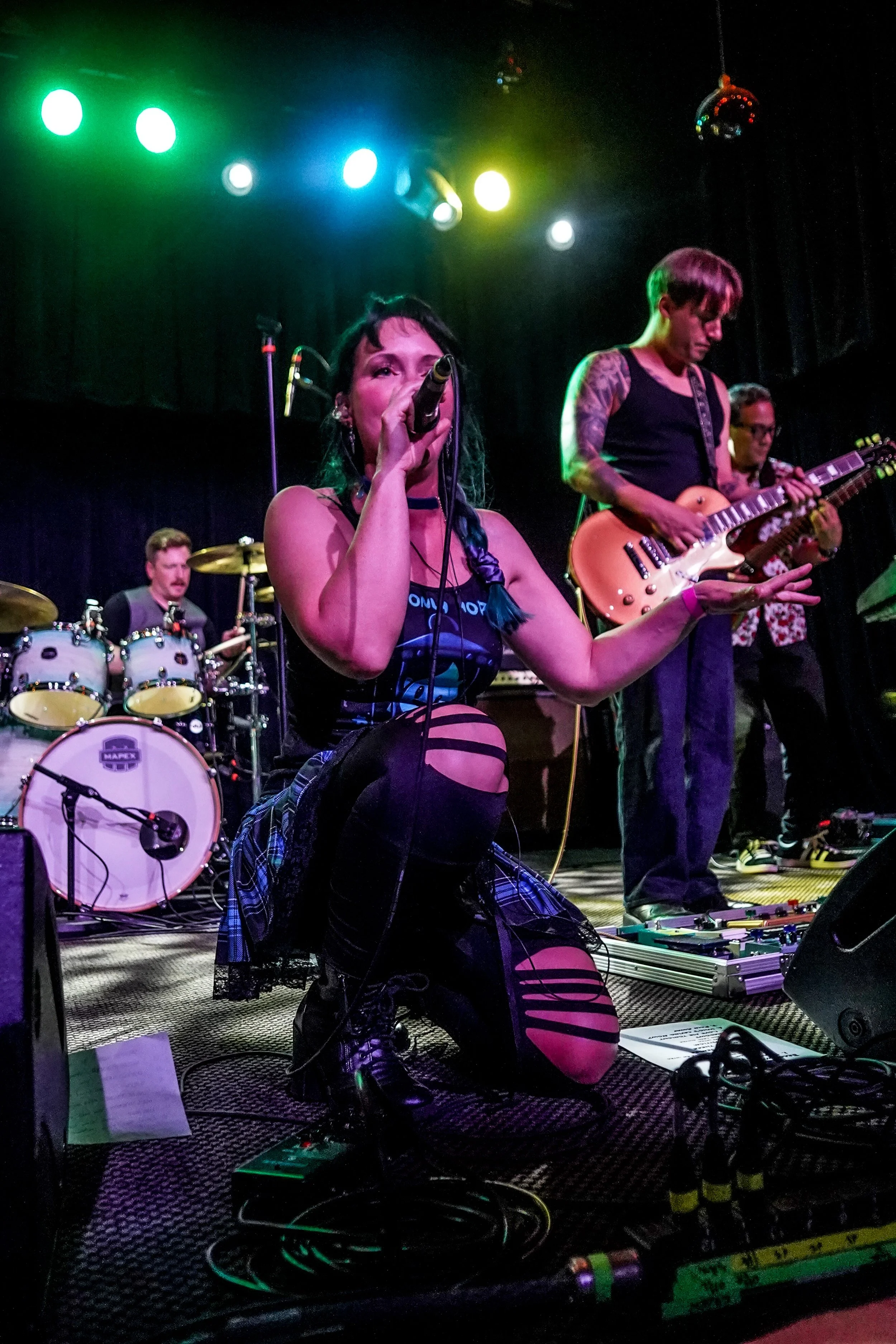Kneeling female vocalist with braid singing into microphone during a live band performance on stage with colored lights, a drummer, and guitarists playing.