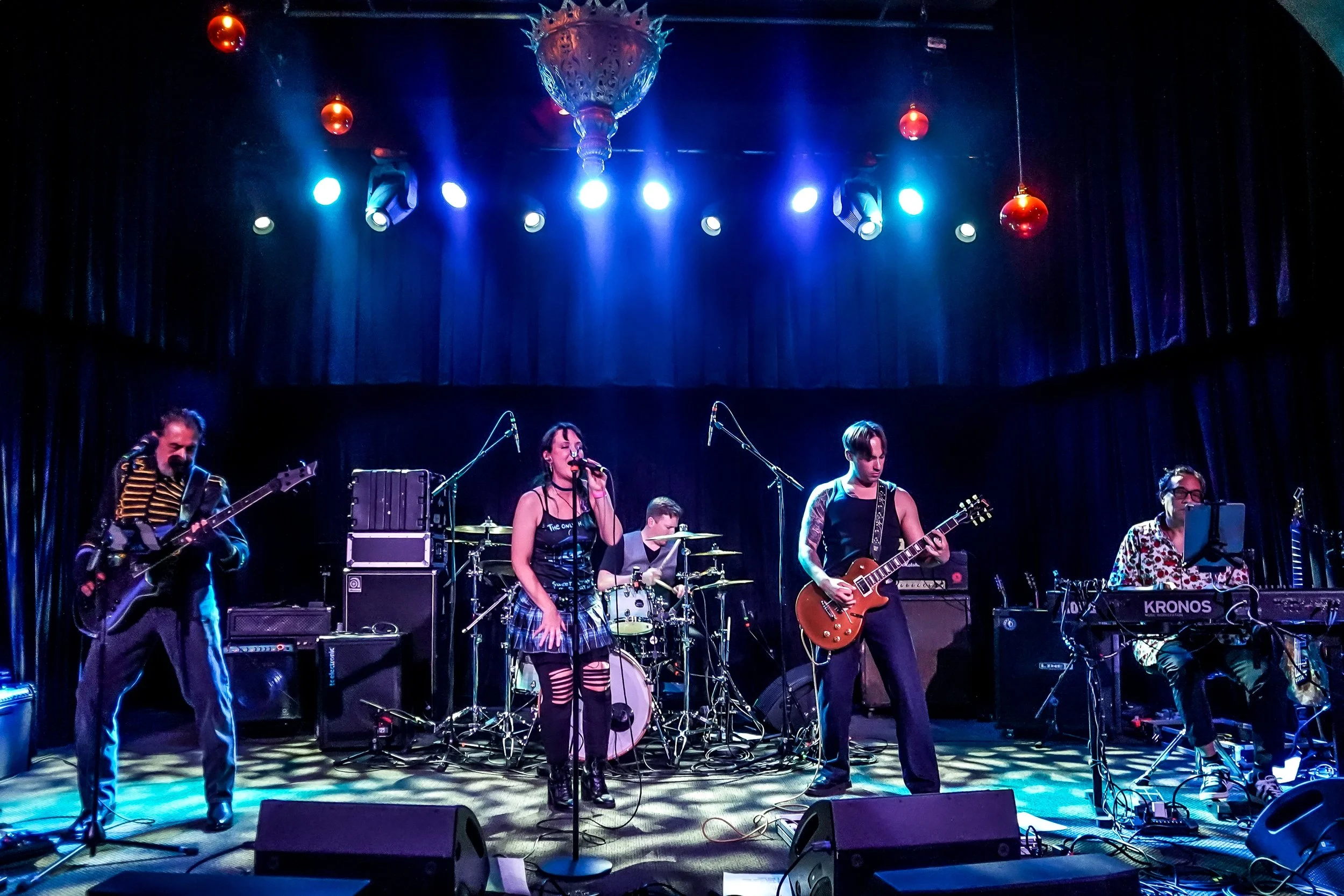 A band performs on stage with colorful lighting and a dark curtain backdrop.