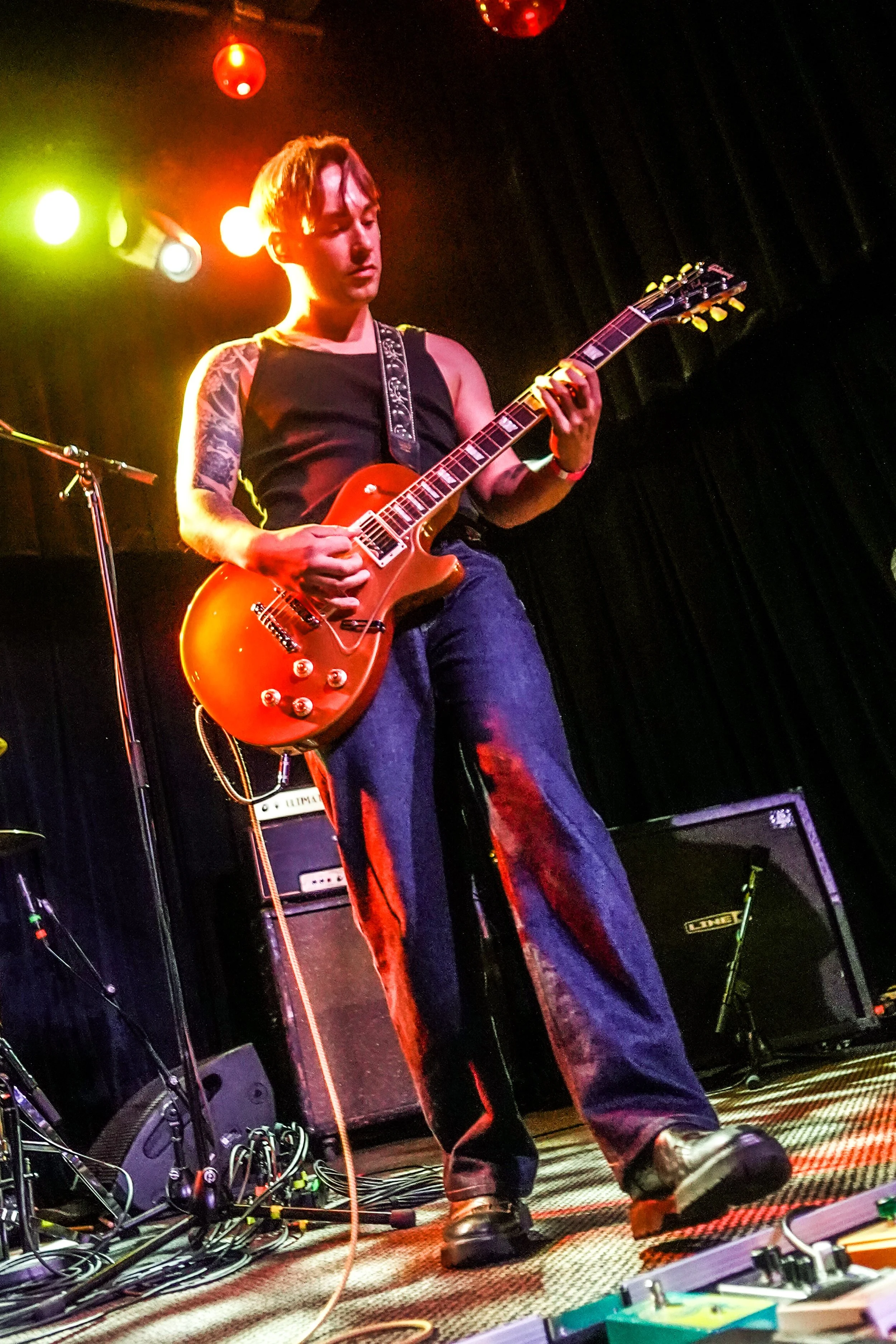 A young man playing an electric guitar on stage, with colorful stage lights and multiple guitar amplifiers behind him.