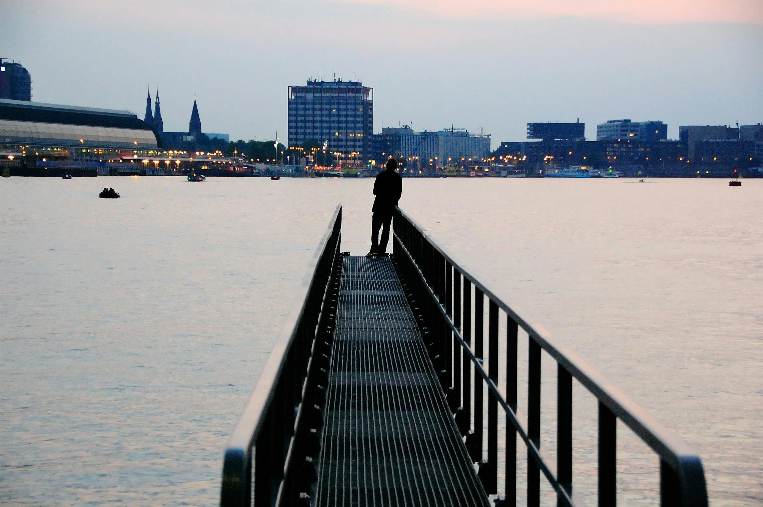 A person standing at the end of a narrow pier overlooking a city skyline and a river, with early evening or dusk lighting.