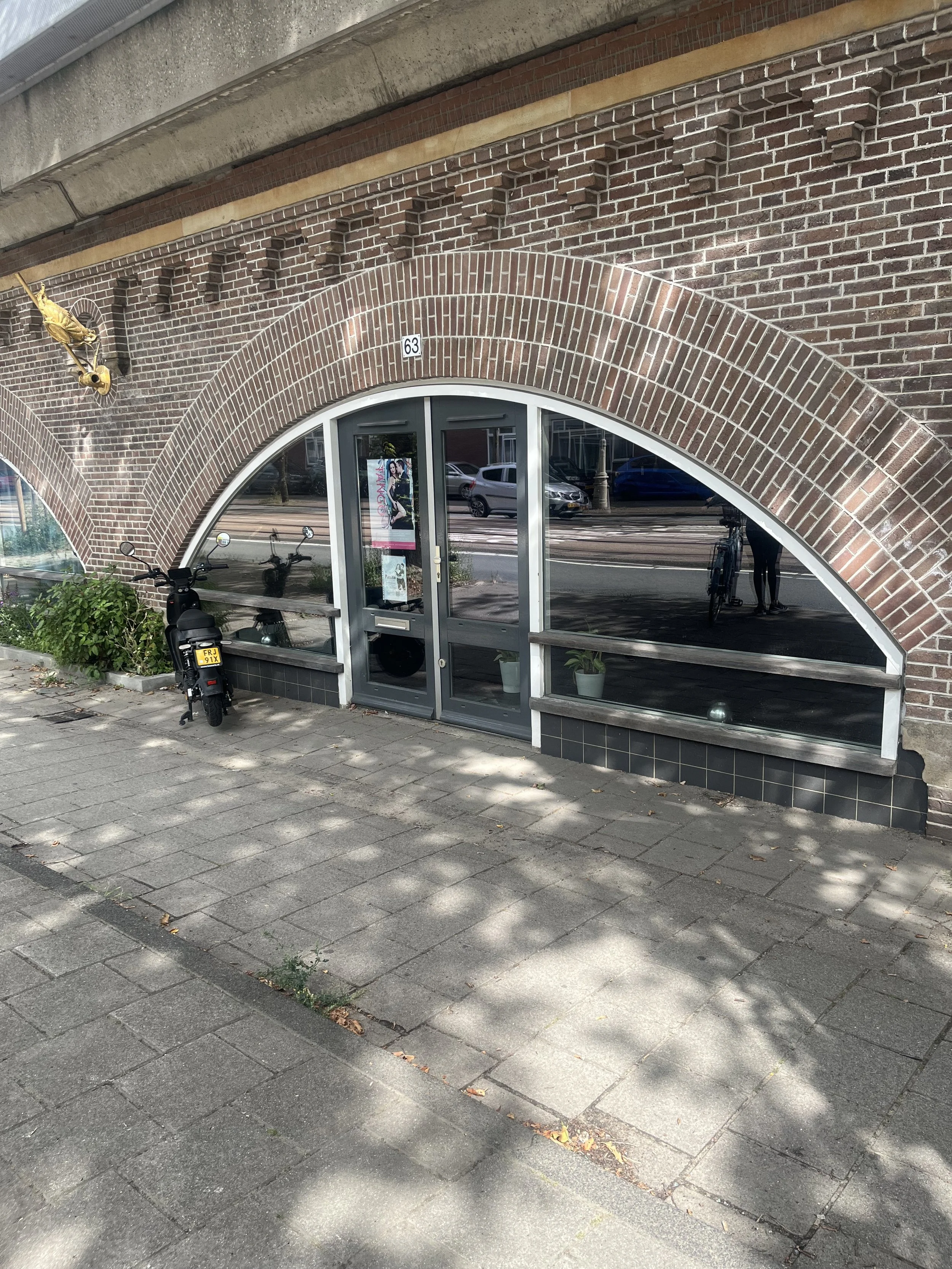 View of a brick building with a rounded arch entrance, glass double doors, a small motorcycle parked outside, and a potted plant inside.