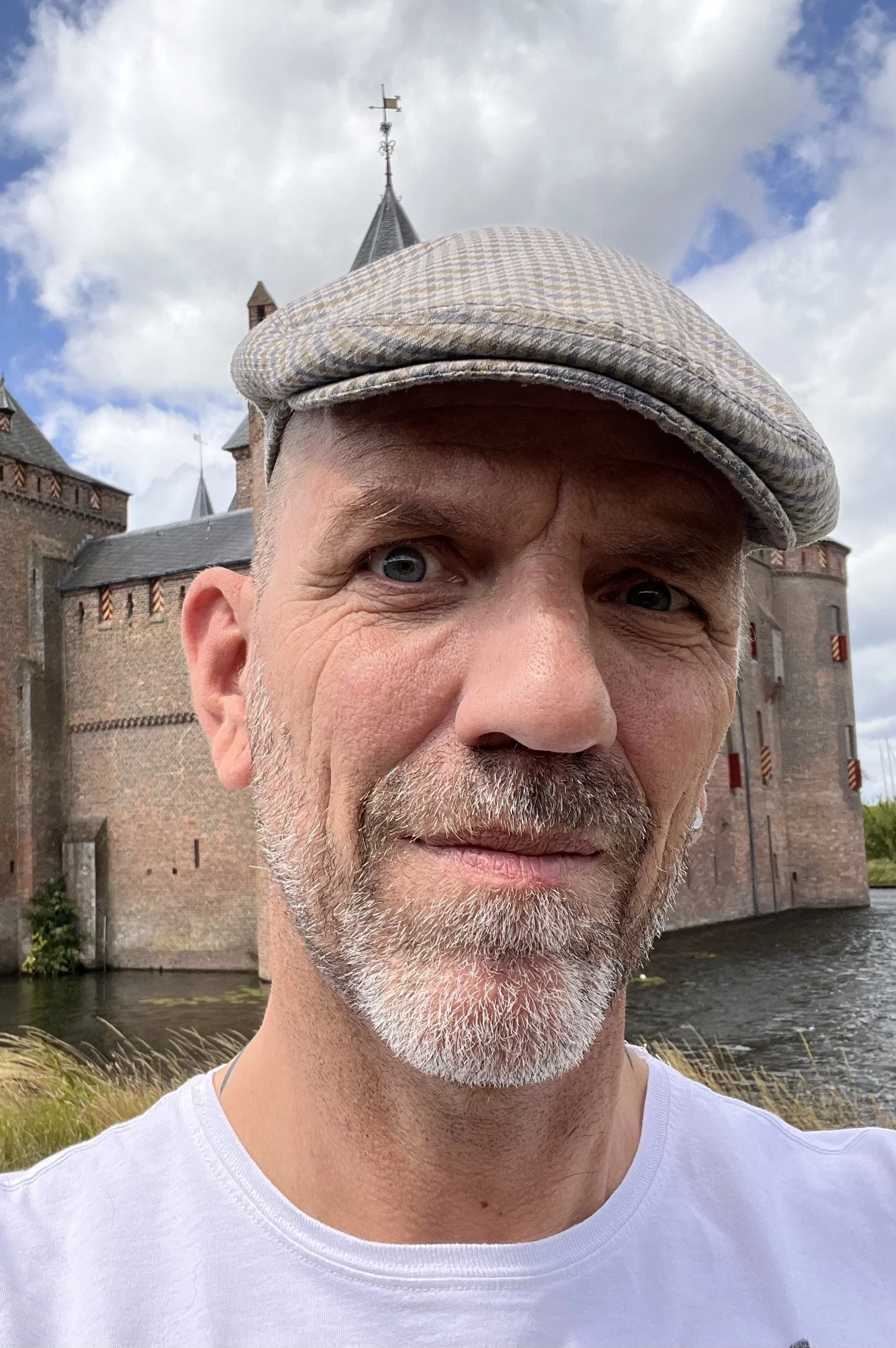 A man with blue eyes, a gray beard, and wearing a flat cap taking a selfie in front of a castle with water and cloudy sky in the background.