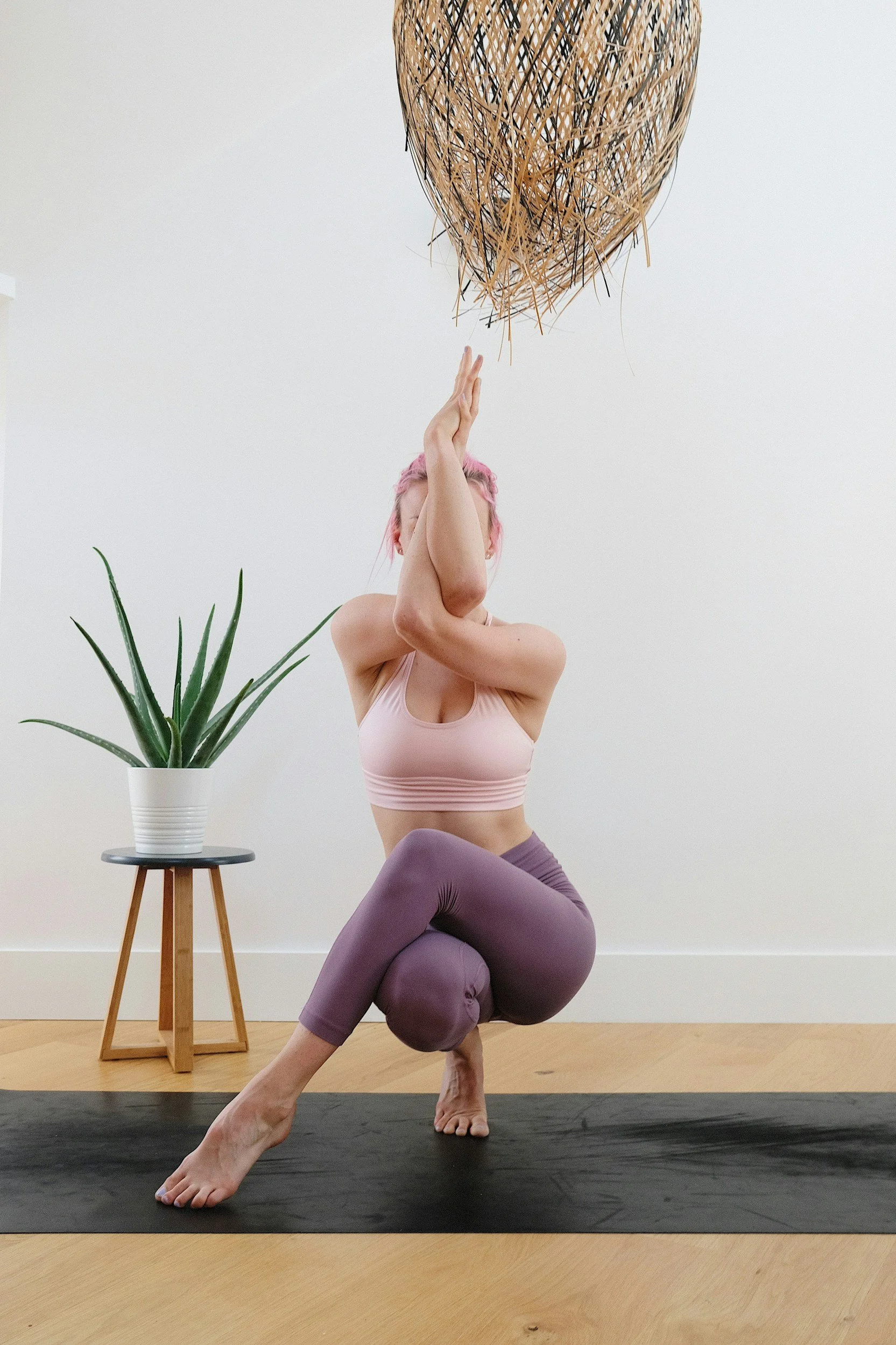 Vrouw oefent yoga in een lichte kamer met houten vloer, een plant op een houten standaard en een decoratieve rieten lamp boven haar hoofd.
