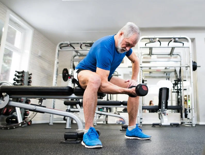 Een oudere man in een fitnessruimte die een dumbbell blijft oefenen op een bank.