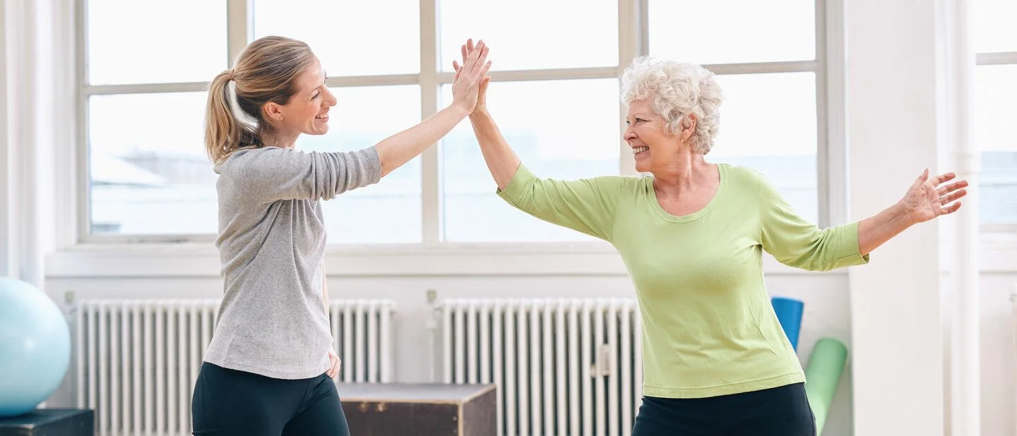 Twee vrouwen geven elkaar een high five in een fitnessruimte met grote ramen.