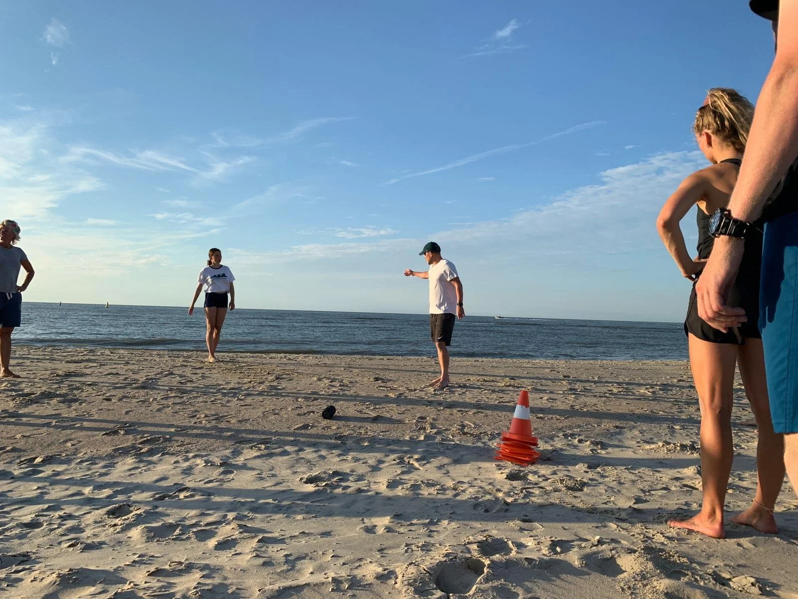 Mensen die strandvoetbal spelen op het strand, met een oranje verkeersstok en zwarte pet op de zandbodem, onder een blauwe hemel.