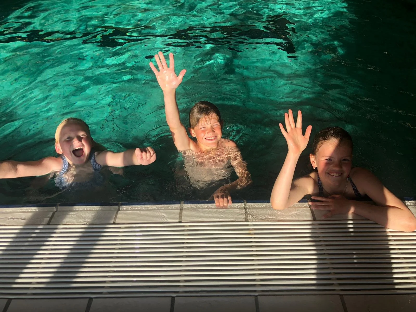 Drie kinderen zwemmen en lachen in het zwembad, met handen omhoog en vriendelijke gezichten.