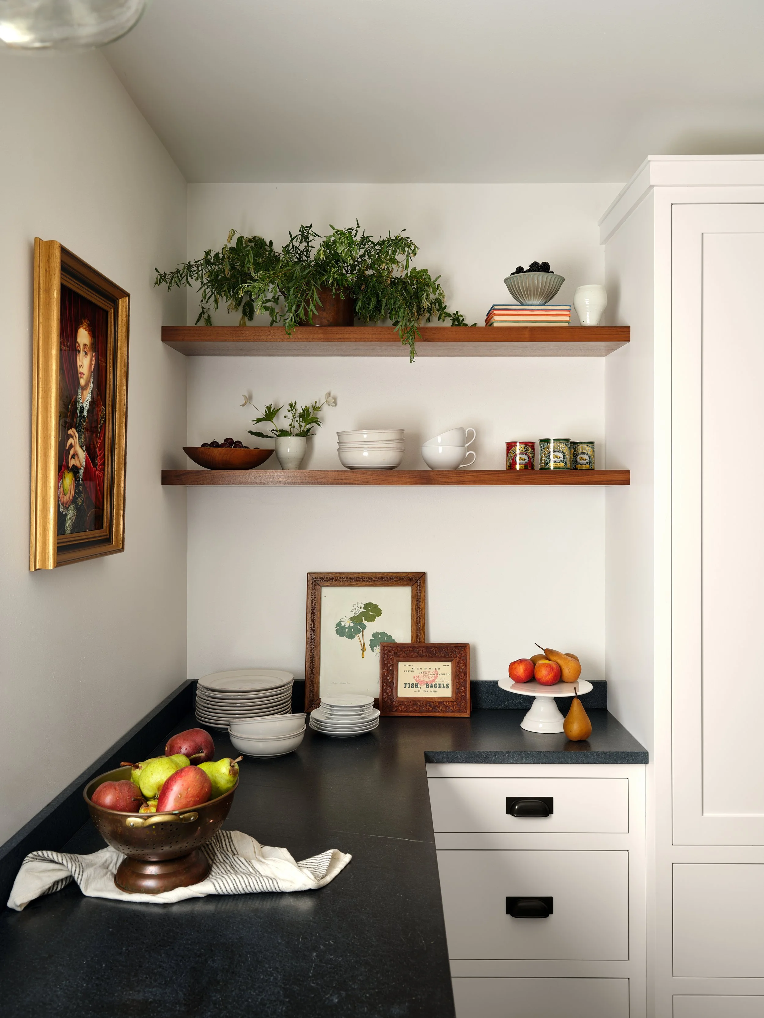 A kitchen corner with white cabinets, two wooden shelves holding plants and bowls, a countertop with apples and framed artwork, and a bowl of apples on a cloth.