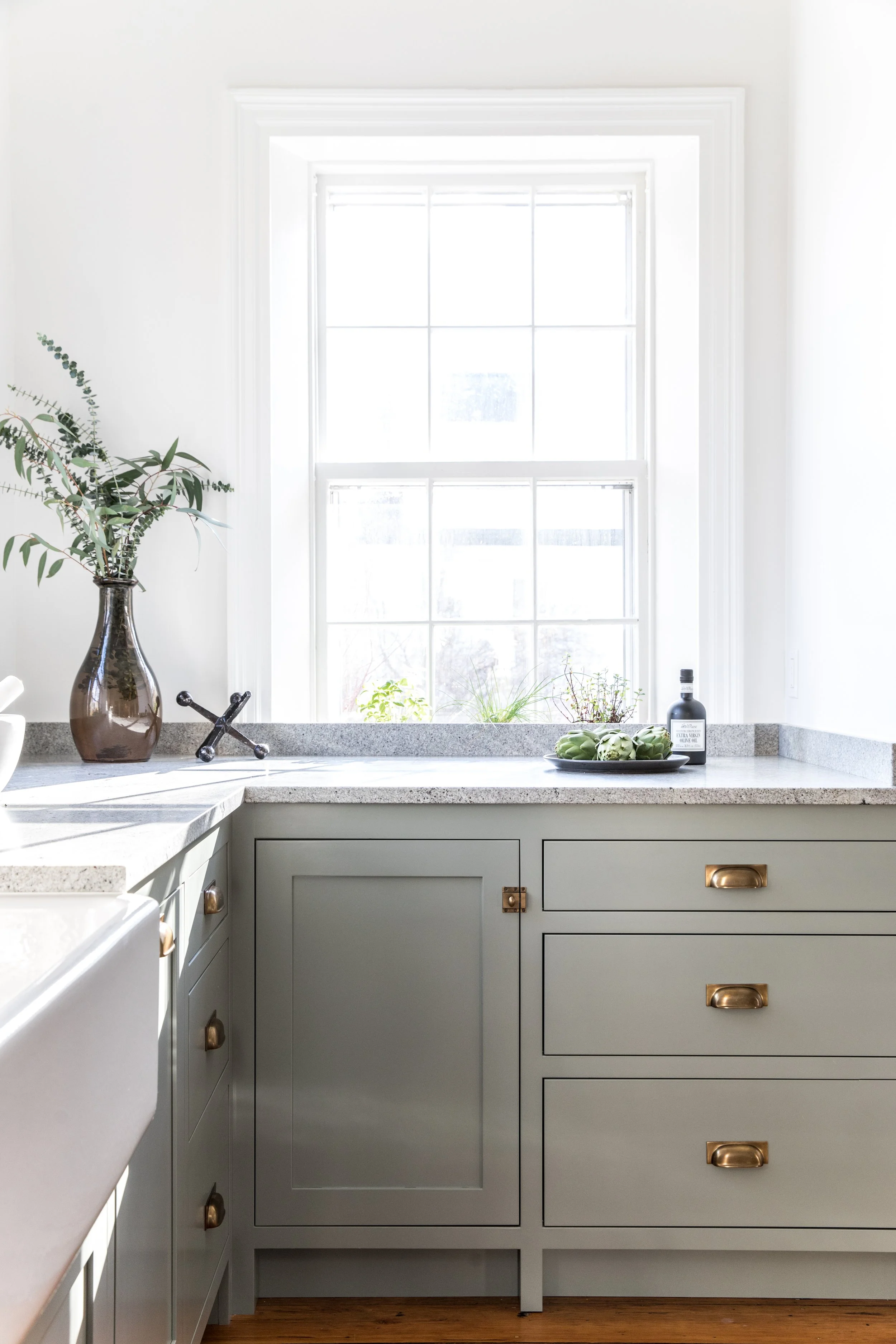 Kitchen with gray cabinetry, a beige countertop, a window with sunlight, a brown vase with leaves, a black faucet, a black bottle, and artichokes on a plate.